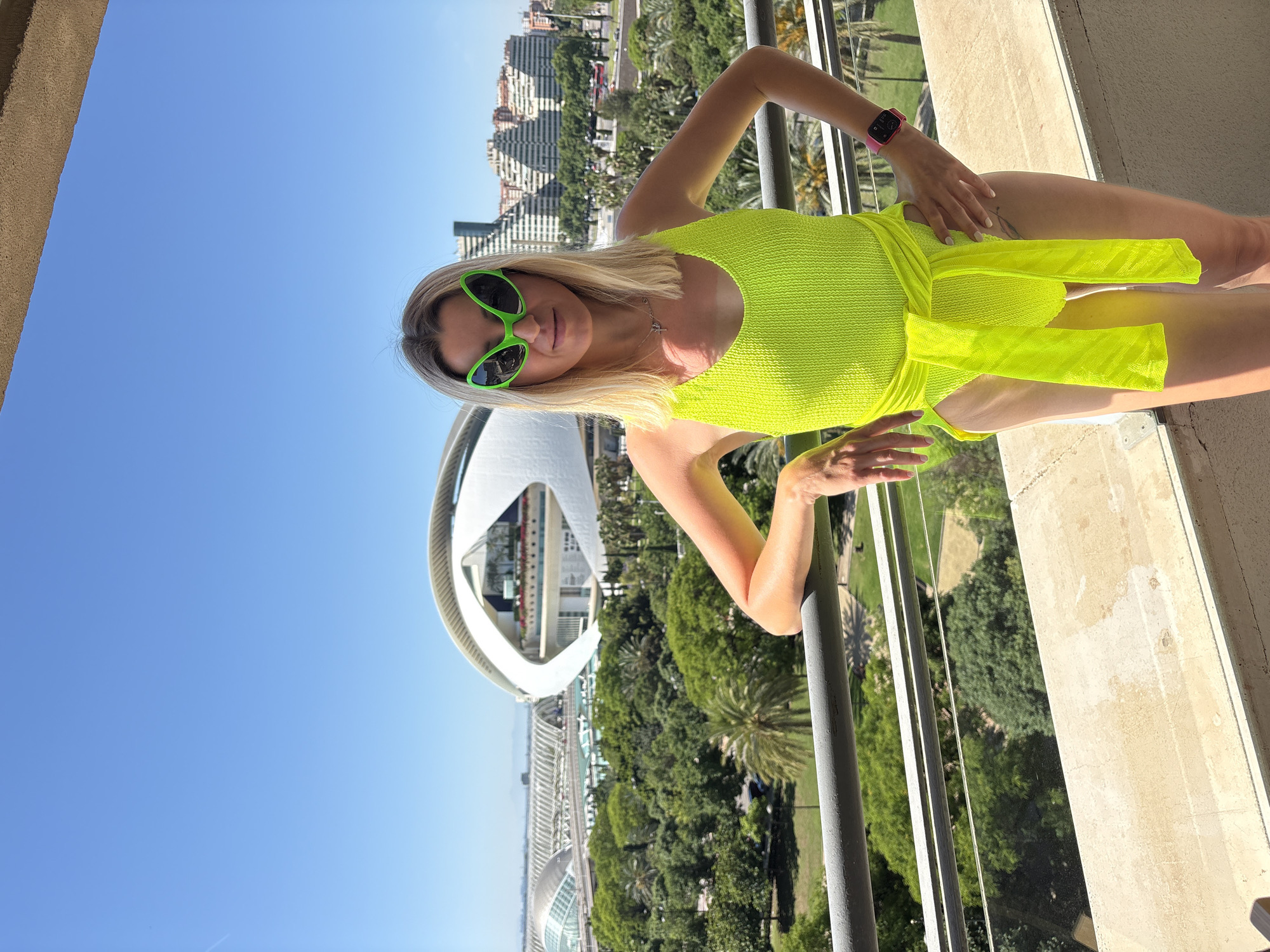 A full-length shot depicts a woman in a neon green outfit and sunglasses, posing on a balcony with the City of Arts and Sciences complex in València, Spain, in the background. She leans against a metal railing, overlooking a lush, green area. The sky is clear and blue. The buildings of the City of Arts and Sciences are visible, with the L'Hemisfèric structure prominently featured.