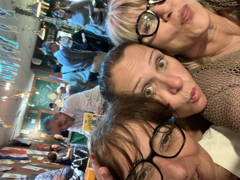 The image displays three adult females in a close-up selfie in the foreground, positioned within an indoor public establishment, likely a bar or pub, in L'Étrat, France.
In the immediate foreground, from left to right:
1. A partially visible adult female with dark hair and black-framed glasses is present on the left. Her right hand, exhibiting red nail polish, is positioned on the head of the central female.
2. A central adult female with dark hair, wide eyes, and puckered lips gazes directly at the camera. She wears a light brown, open-knit garment.
3. A partially visible adult female on the right, with blonde hair and black-framed glasses, also has puckered lips and looks toward the camera. She wears a light brown, open-knit garment and a delicate necklace.
The background shows a populated commercial space characterized by wood-paneled walls, sections painted green, and numerous flags or pennants strung from the ceiling and affixed to walls. A "WC" sign is visible on a green wall, and a dartboard is mounted nearby. Artificial lighting illuminates the area. Multiple adult patrons are present, out of focus. A man with graying hair, wearing a light-colored long-sleeve shirt, is visible in the midground, looking down at a book with a green cover titled "ST PATRICK." Other individuals are standing and conversing throughout the venue, some near tables or a bar counter. A black bag rests on a surface in the left midground, and a yellow bowl is partially visible in the far left foreground. The scene depicts a social gathering inside a public house.