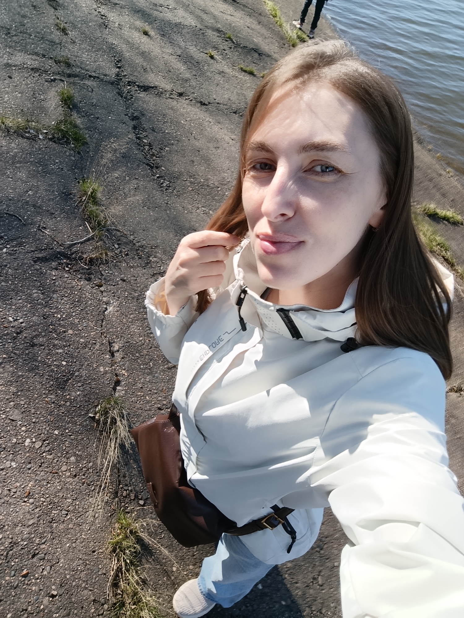 A young, fair-skinned woman is taking a selfie outdoors. She has long brown hair and blue eyes. She is wearing a white hooded jacket, light blue jeans, and white sneakers. A brown leather bag is slung across her body.
She is standing on a paved surface next to a body of water. The paving is cracked and has grass and weeds growing in the cracks. The water's edge is visible in the upper right of the frame. In the upper background, someone’s legs can be seen.
The setting appears to be along a waterfront or embankment in an urban area, possibly Chaikovsky, Russia.