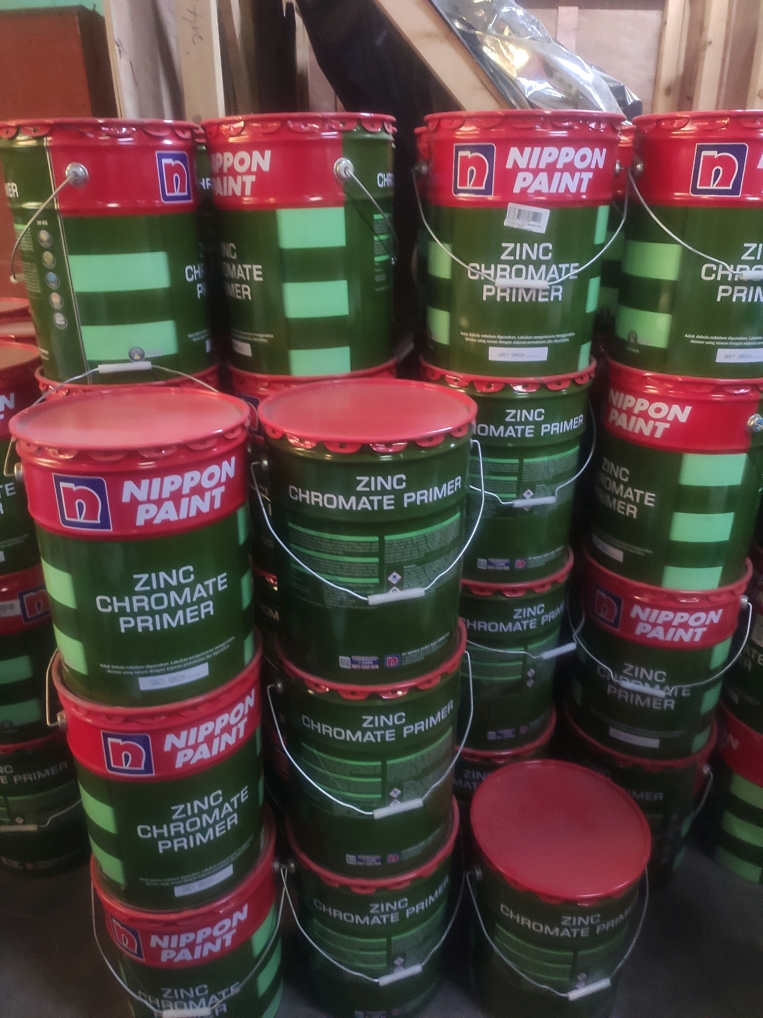A tall, eye-level, medium shot captures a cluttered stack of green and red paint cans from Nippon Paint. The cans are cylindrical with red rims and lids, stacked unevenly in several columns. The prominent text on the cans reads "NIPPON PAINT" and "ZINC CHROMATE PRIMER". Some cans are fully visible, while others are partially obscured by the stack. The background is dark and out of focus, suggesting an indoor setting, possibly a warehouse or storage area. The lighting is dim, casting shadows that add to the stacked and cluttered appearance of the scene. The overall impression is one of a commercial storage of paint products.