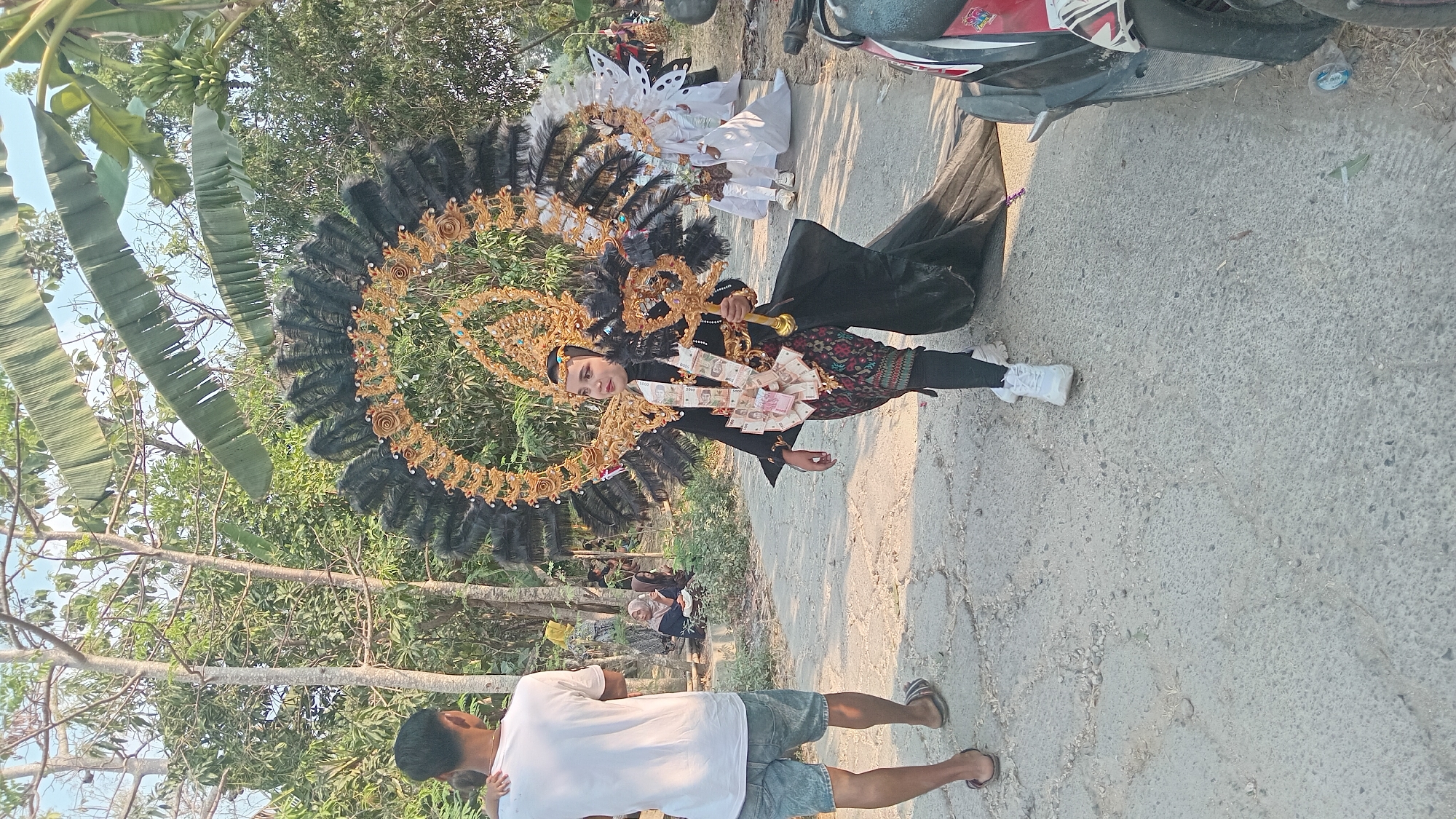 In Kedungwaru, Indonesia, a young woman walks along a paved road, adorned in an elaborate, traditional costume. Her outfit features a large, ornate headdress made of black feathers and gold accents, a black long-sleeved top, and a patterned skirt. She carries a golden scepter and has stacks of Indonesian Rupiah banknotes attached to her chest. Behind her, other people are visible in the background, including a person in a white costume and another wearing a headscarf. The setting appears to be outdoors, with trees and vegetation surrounding the area, suggesting a festive or ceremonial event. A motorcycle is parked on the right side of the road.
