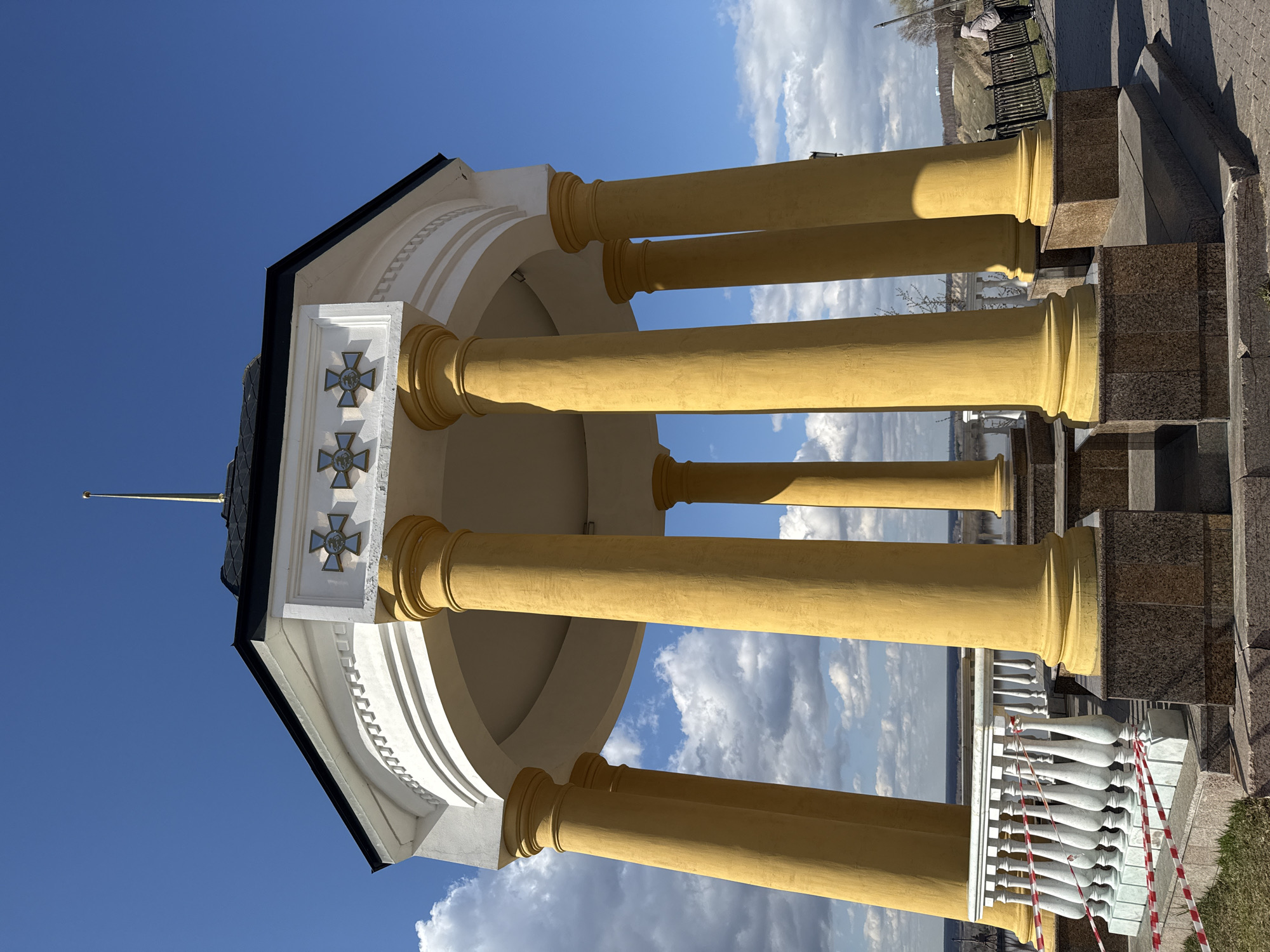 This media file captures a classical-style rotunda or gazebo, likely situated at a scenic overlook. The structure features several prominent, golden-yellow columns with ornate bases and capitals, supporting a white entablature. The entablature has decorative white panels, each adorned with a grey or silver cross emblem. A dark, possibly grey or black, polygonal roof tops the structure, crowned by a golden, pointed spire or finial.
At the base of the rotunda, a white balustrade with elegantly shaped balusters is partially visible, with sections cordoned off by red and white striped warning tape. The foundation and pathway immediately surrounding the structure are constructed from dark granite blocks.
The scene is set under a bright blue sky with scattered white cumulus clouds, indicating sunny weather, likely late morning or early afternoon given the clear light and shadows cast by the columns. In the background, a panoramic landscape extends, revealing what appears to be a wide river or body of water, flanked by distant green banks and trees. A solitary individual, wearing a light-colored jacket, is visible in the far background, seemingly sitting or standing on a bench or railing, observing the view. No specific activity is occurring. The image conveys a sense of peaceful grandeur. There is no visible text in the image. The location context for this structure is Tobolsk, Russia.
