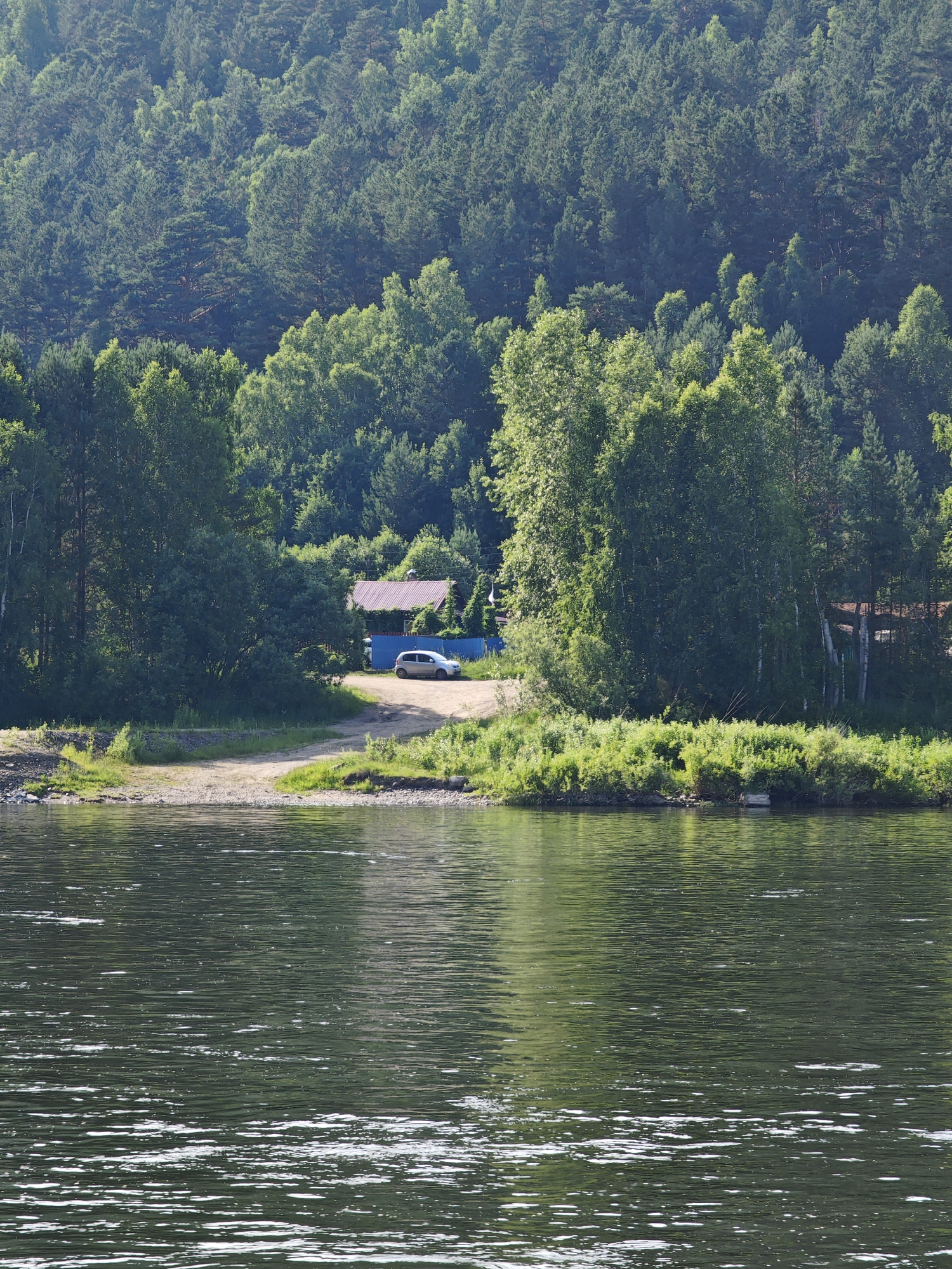 This image depicts a serene waterside scene in Pristan', Russia, with a dense, green forest as the backdrop. In the foreground, the dark, rippling water of a river dominates the lower half of the frame, reflecting the surrounding trees and sky. The riverbank is lined with lush green vegetation and a gravelly shore. Further inland, a dirt road leads to a small clearing where a silver car is parked. Behind the car, a building with a reddish-brown roof is visible, nestled amongst the trees. The forest itself is a thick expanse of various trees, predominantly coniferous, with lighter green deciduous trees interspersed. The overall impression is one of natural beauty and tranquility, with a hint of human presence.