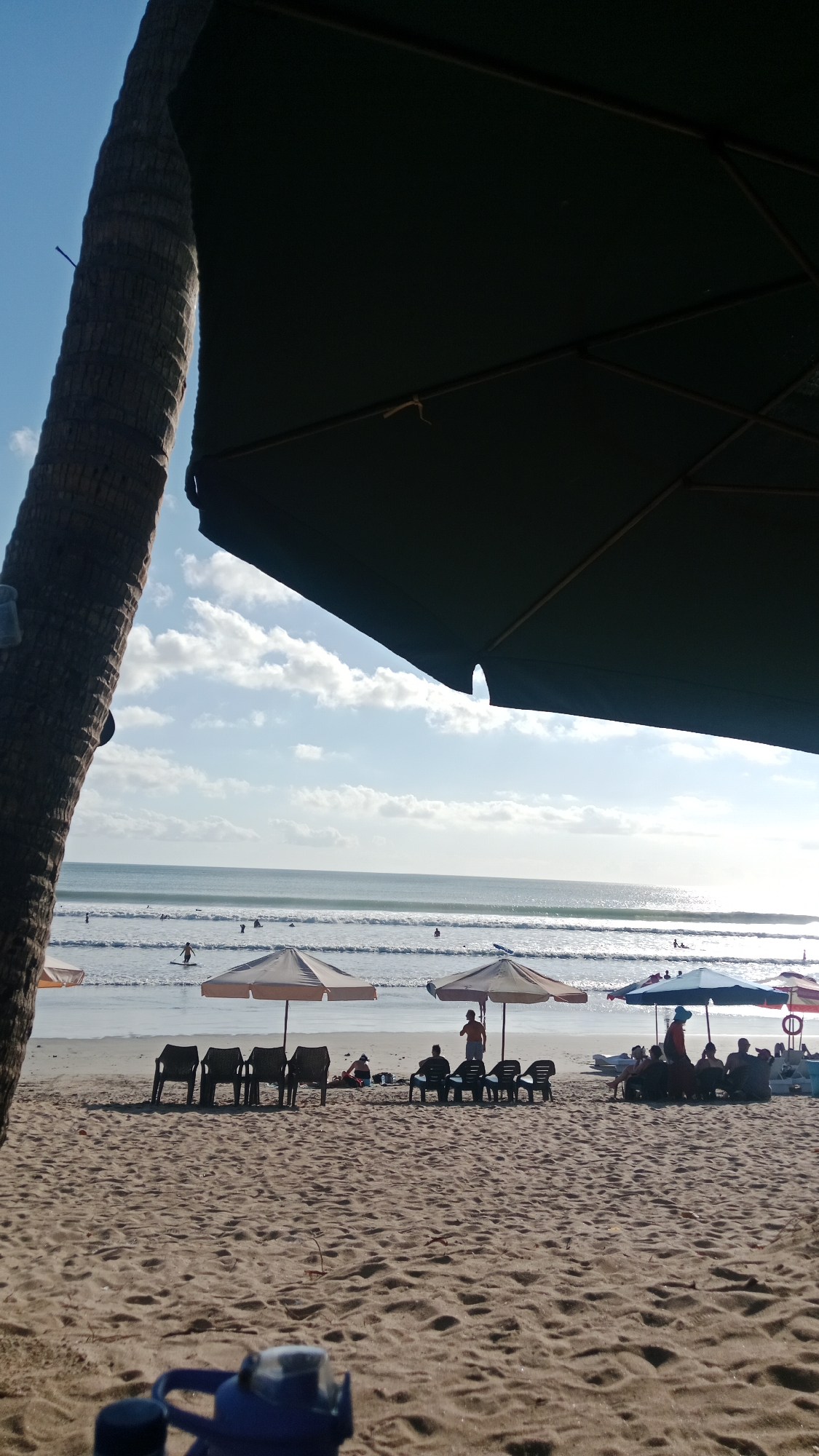 A wide-angle shot captures a sandy beach in Kuta, Indonesia, under a bright, partly cloudy sky. In the foreground, a large, dark umbrella partially obscures the view, with a portion of a thick, textured palm tree trunk visible on the left. The beach is dotted with numerous empty, dark-colored plastic chairs arranged in rows, some partially shaded by various beach umbrellas. Further back, a few people are seated under umbrellas, and others are scattered on the sand. The ocean is visible in the mid-ground, with gentle waves breaking on the shore. Several individuals are in the water, some appearing to be swimming or surfing, with a lone figure standing in the shallow water. The sunlight reflects brightly off the ocean's surface. The scene depicts a relaxed atmosphere on a typical beach day in Kuta.