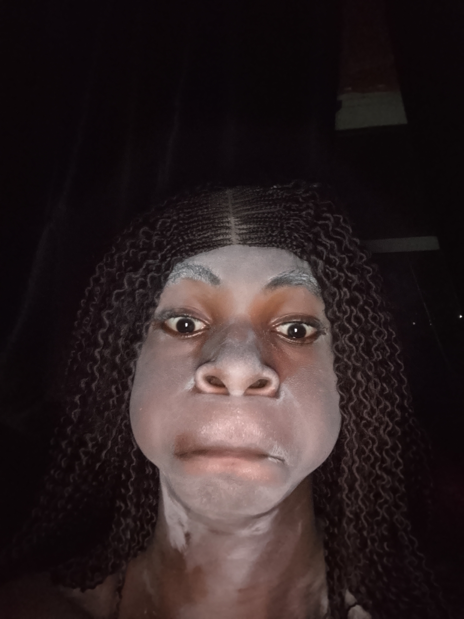 A close-up, dimly lit portrait features an individual, likely female, with a playful or exaggerated expression. The person has long, dark, intricately braided hair that frames their face. Their skin appears to be covered in a light, whitish substance, resembling a face mask or powder, particularly noticeable on the forehead, nose, cheeks, and chin. Their eyes are wide open and looking directly at the viewer, while their cheeks are puffed out, and their mouth is pursed in a slight frown. The background is extremely dark and indistinct, suggesting low light conditions or an indoor setting with minimal illumination. There are faint hints of vertical lines or textures in the upper background and a dark, rectangular shape on the right side in one of the crops, but these details are not clear enough to identify a specific location. The lighting seems to come directly from the front, highlighting the person's face against the dark surroundings. No text is visible in the image.