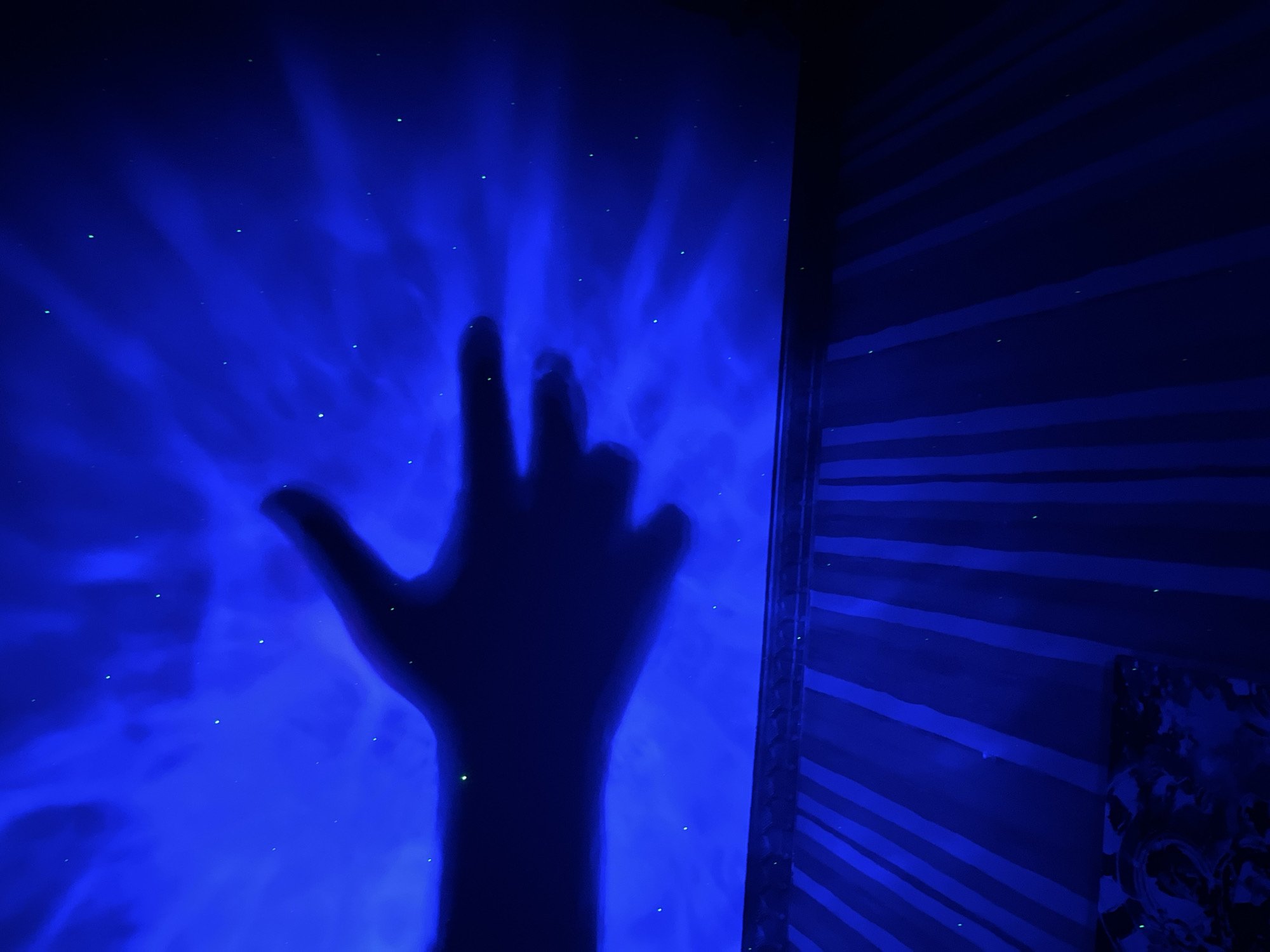 A silhouette of a hand, possibly a child's due to its size, is cast against a blue background illuminated by a projector that creates swirling cloud-like patterns and faint green specks resembling stars. The hand is positioned horizontally, with the thumb extended and the fingers slightly curled. Below the projected image, a striped surface, likely a rug or blanket, is visible with alternating dark and light blue stripes. A portion of a patterned fabric is also visible in the lower left corner. The overall scene suggests a playful or artistic setup, possibly in a bedroom or dimly lit room. There is no information to indicate the location in Saint Petersburg, Russia.