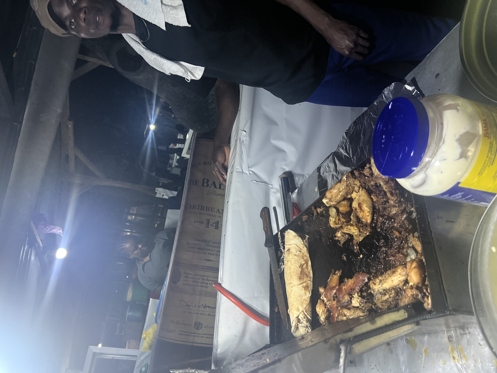 An overhead, tilted image captures a street food vendor preparing food on a grill at night in Abuja, Nigeria. The primary subject is a man, likely the vendor, looking up towards the camera. He wears a dark shirt, blue trousers, and a light-colored headwrap or towel. A white cloth is draped over his shoulder.
He stands behind a white-covered counter where a metal grill or hot plate is positioned. On the grill, pieces of cooked meat, resembling chicken, are visible, along with a wrapped item, possibly a shawarma or similar flatbread wrap. Two knives are placed near the food. A bright orange gas hose is also visible on the counter, likely connected to the grill.
To the right of the grill, a large plastic jar of mayonnaise with a blue lid is prominently displayed, with the brand name "BAMA" visible on its yellow label. Partially obscured text below it reads "SOYBEAN OR WA".
In the background, a large cardboard box for "THE BALVENIE" Distillery Caribbean Extra Matured whiskey, "AGED 14" years, is stacked. Further text on the box mentions "SPECIALLY SELECTED," "David Stewart," and "THE BALVENIE MALT MASTER," along with location details like "BALVENIE MALTINGS, DUFFTOWN, BANFFSHIRE, SCOTLAND AB4". Another person is faintly visible in the dark background. The scene is illuminated by artificial lights, creating bright flares and dark shadows, typical of a nighttime outdoor market or food stall.