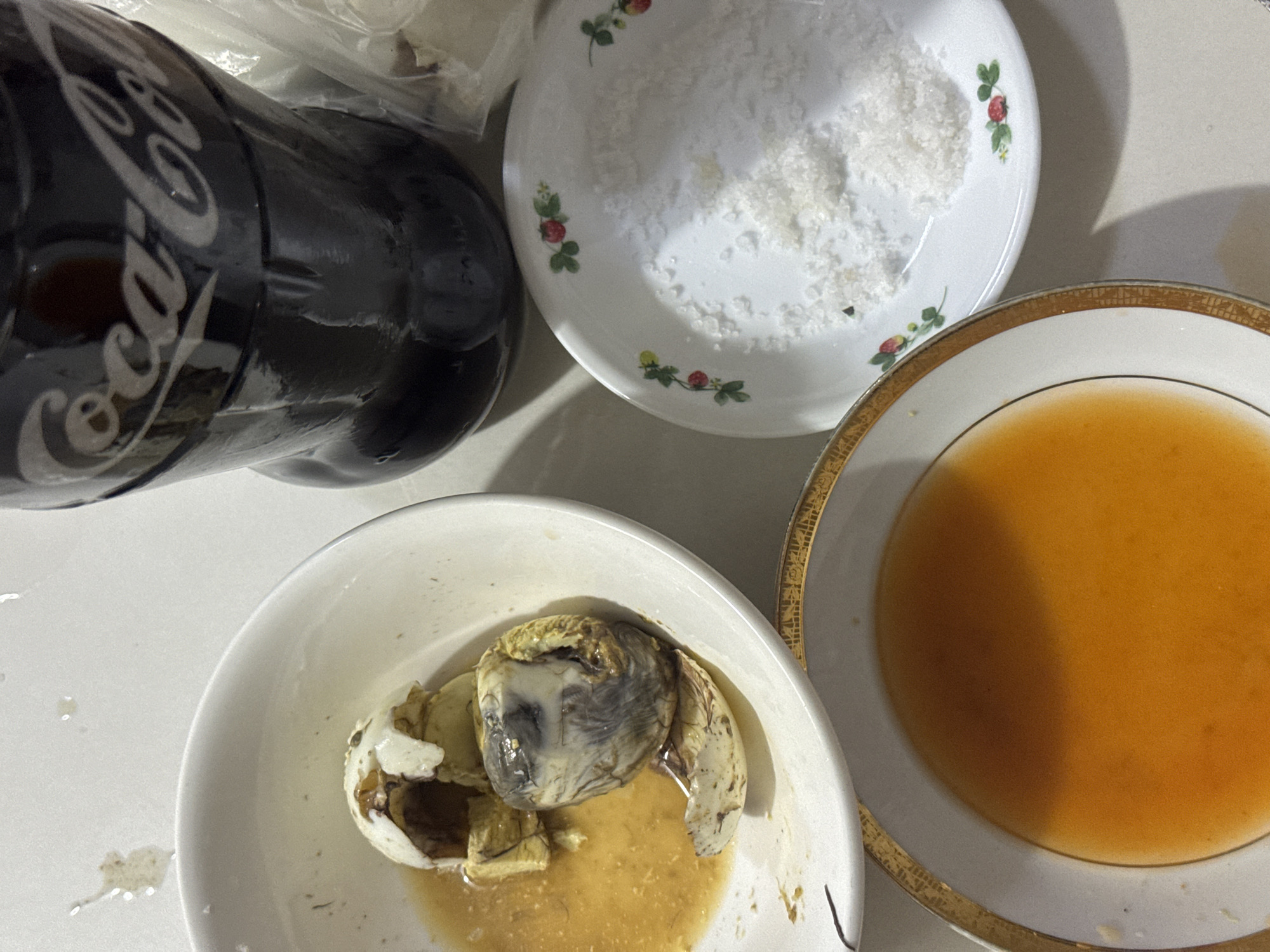 An overhead shot presents a typical Filipino snack setup on a light-colored surface, likely a table or counter, in Davao City, Philippines. The central subject is a balut, a fertilized duck embryo, partially opened and resting in a white bowl filled with a yellowish-brown broth. To the right of the balut, a white bowl with an ornate gold rim holds an orange-brown liquid, likely a dipping sauce or vinegar. Above these, a small white plate, decorated with red strawberries and green leaves, contains a pile of coarse white salt. In the upper left corner, a black Coca-Cola bottle, displaying the iconic "Coca-Cola" text in white script and showing condensation, lies horizontally, suggesting it is a cold beverage. The lighting casts clear shadows, particularly across the bowls, indicating an indoor setting, possibly under artificial light. The table surface also shows a few minor splatters or marks.