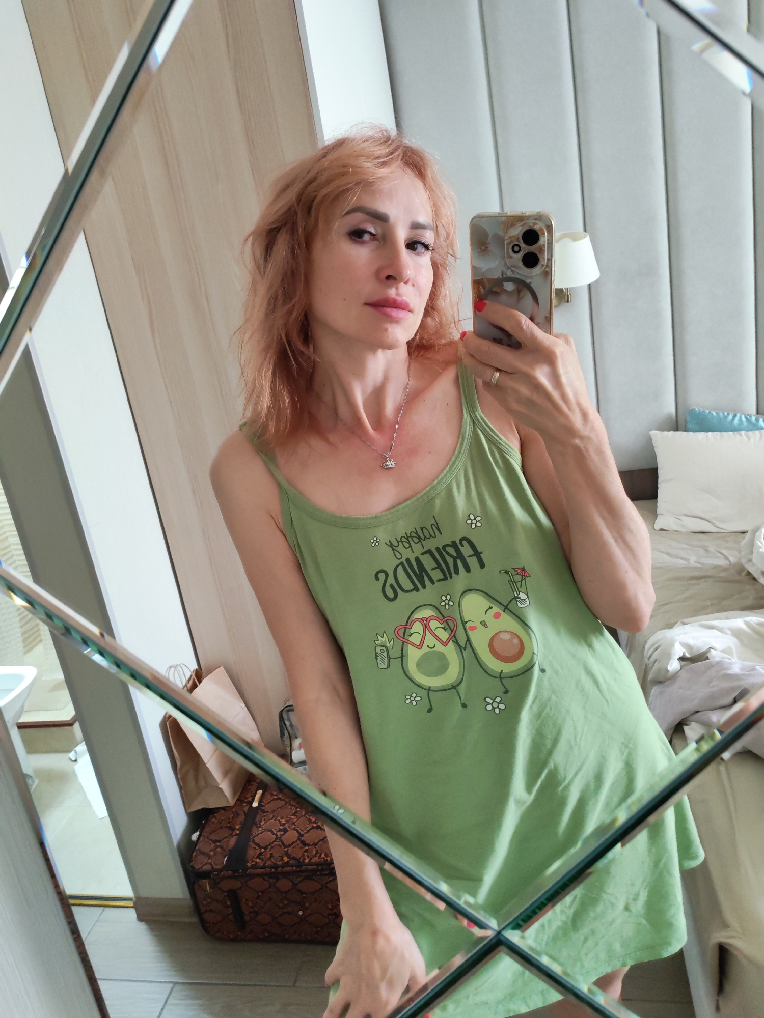 The image shows a woman taking a mirror selfie. She has light red hair, is wearing a light green tank top with a graphic of two avocados and the text "FRIENDS" printed in reverse, and a necklace. She is wearing a ring on her right hand. Her left arm is down by her side and her right arm is holding up her phone to take the picture. The background of the photo shows a bedroom with a large headboard, pillows, and a lamp. To the left of the woman, a large mirrored closet door is visible, which is reflecting the woman and the room. A brown patterned suitcase and a paper bag are on the floor in front of the closet. A white sink and part of a tiled wall are visible in the far left of the reflection. The lighting suggests it is daytime. The setting appears to be a hotel room or bedroom.