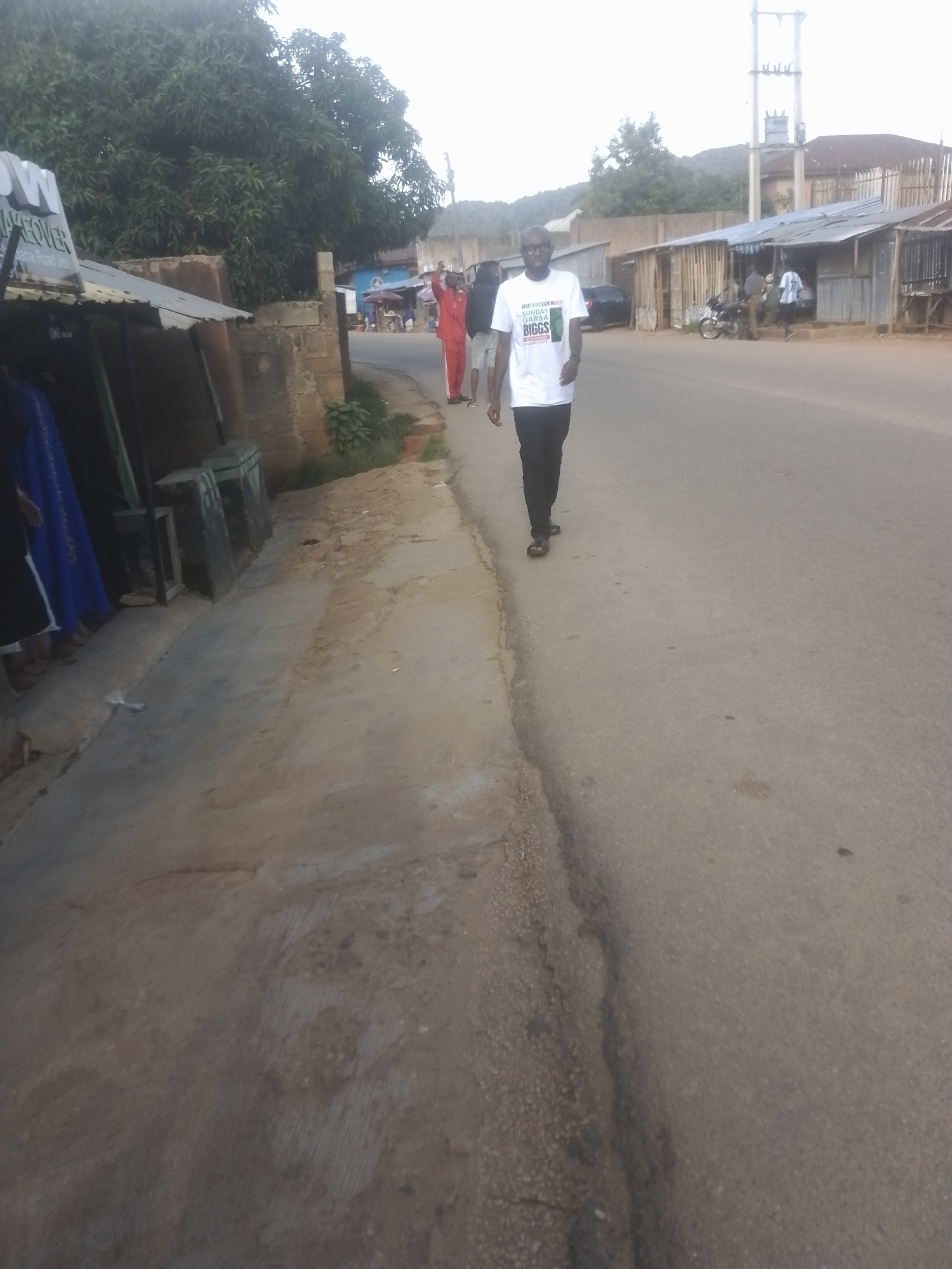 A man wearing glasses and a white t-shirt with text and black pants walks on a paved road in what appears to be an outdoor marketplace or a developing area. The t-shirt has green text that reads "BIGGS" and other words in a smaller font above and below. The man is walking from left to right across the image, facing slightly towards the viewer. To the left of the road, there are several makeshift structures with corrugated metal roofs, some appearing to be shops or stalls. A few people, some wearing traditional clothing, are visible near these structures. Behind the man, more buildings and trees are visible, and a power pole with electrical equipment stands in the background. The road is asphalt, and the area appears to be somewhat dusty, suggesting a warm, dry climate. The overall lighting suggests it might be late afternoon or early morning.