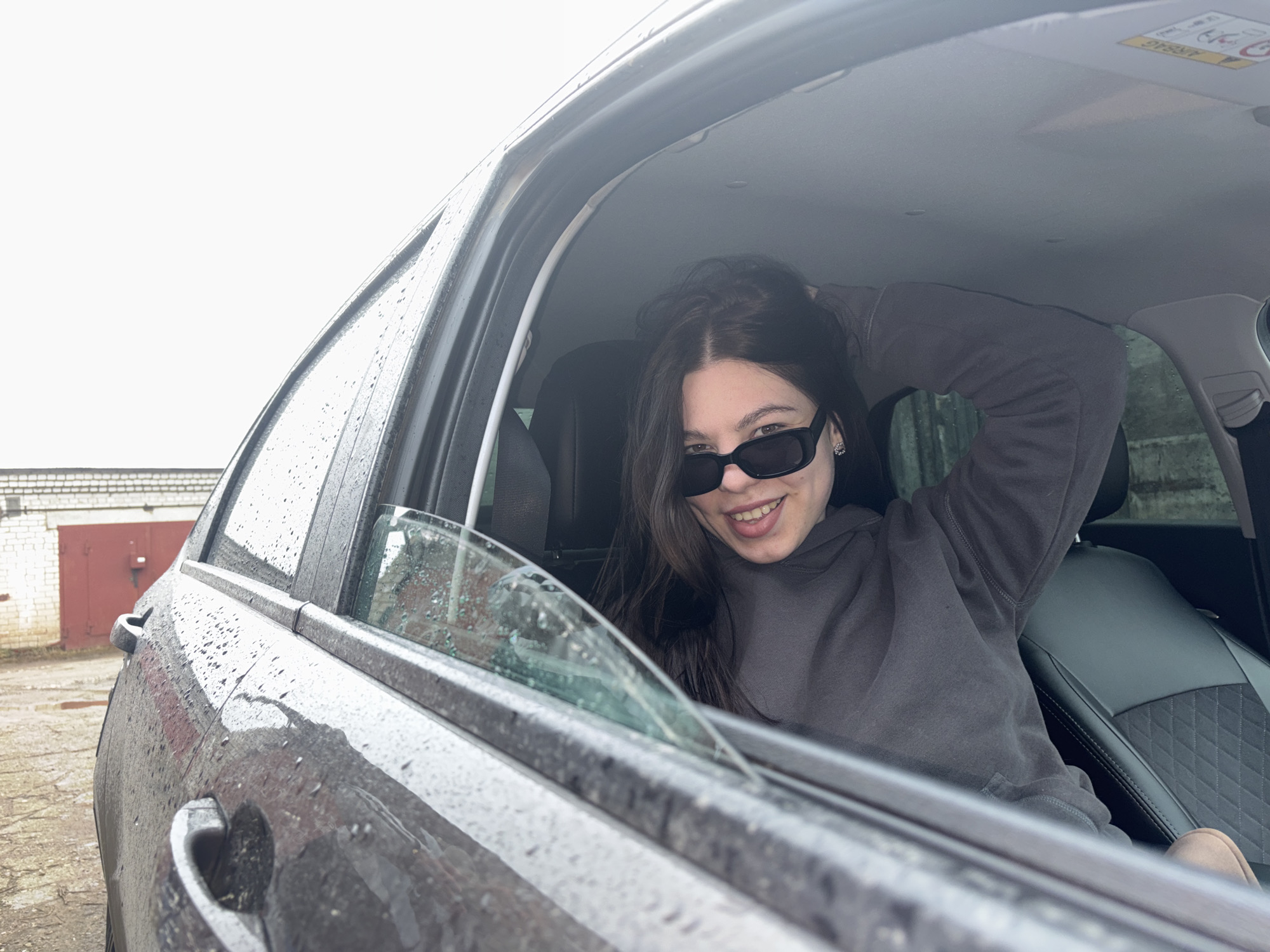A young woman wearing sunglasses is in the driver's seat of a car, with the window partially open. She is smiling and has her hand in her hair. The car is parked outside in a location that appears to be in Perm, Russia, judging by the style of the buildings and general appearance of the surrounding environment. The car is wet, indicating it has been raining or is currently raining. The building in the background appears to be a garage or outbuilding with a red door.