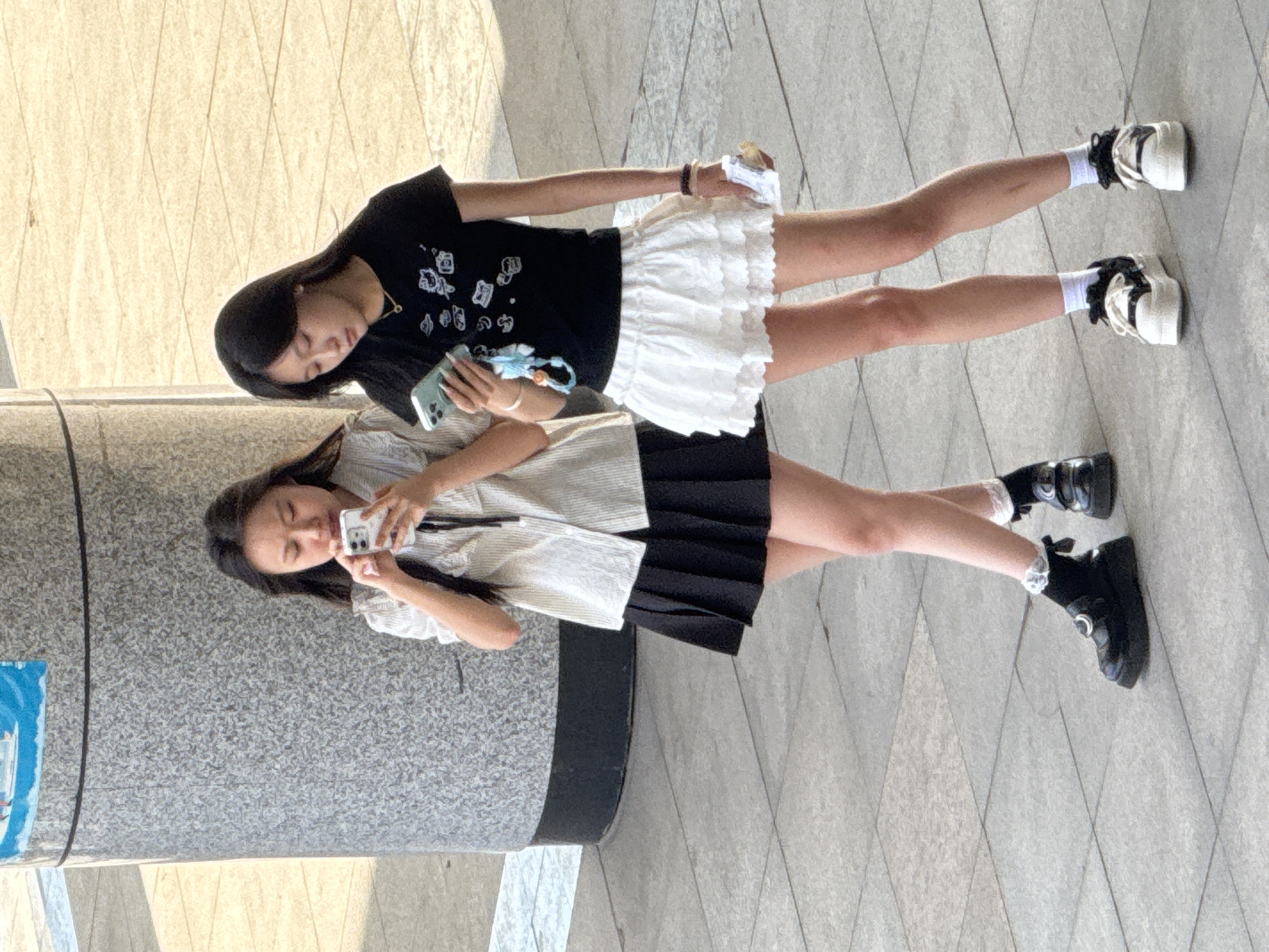 Two young women are walking on a paved sidewalk in front of a large, cylindrical, stone-clad column. The woman on the left is wearing a light-colored, vertically striped shirt with a ruffled collar and a black pleated skirt. She is holding a white smartphone in her hands and looking down at it. She is also wearing white socks with lace trim and black platform shoes. The woman on the right is wearing a black t-shirt with a graphic print and a white ruffled skirt. She is holding a light blue-green smartphone in her left hand and looking down at it. She is also wearing white socks and white platform sneakers with black accents. The ground is paved with light gray square tiles. The lighting suggests it is daytime. The overall scene depicts a casual moment between two individuals in an urban environment.