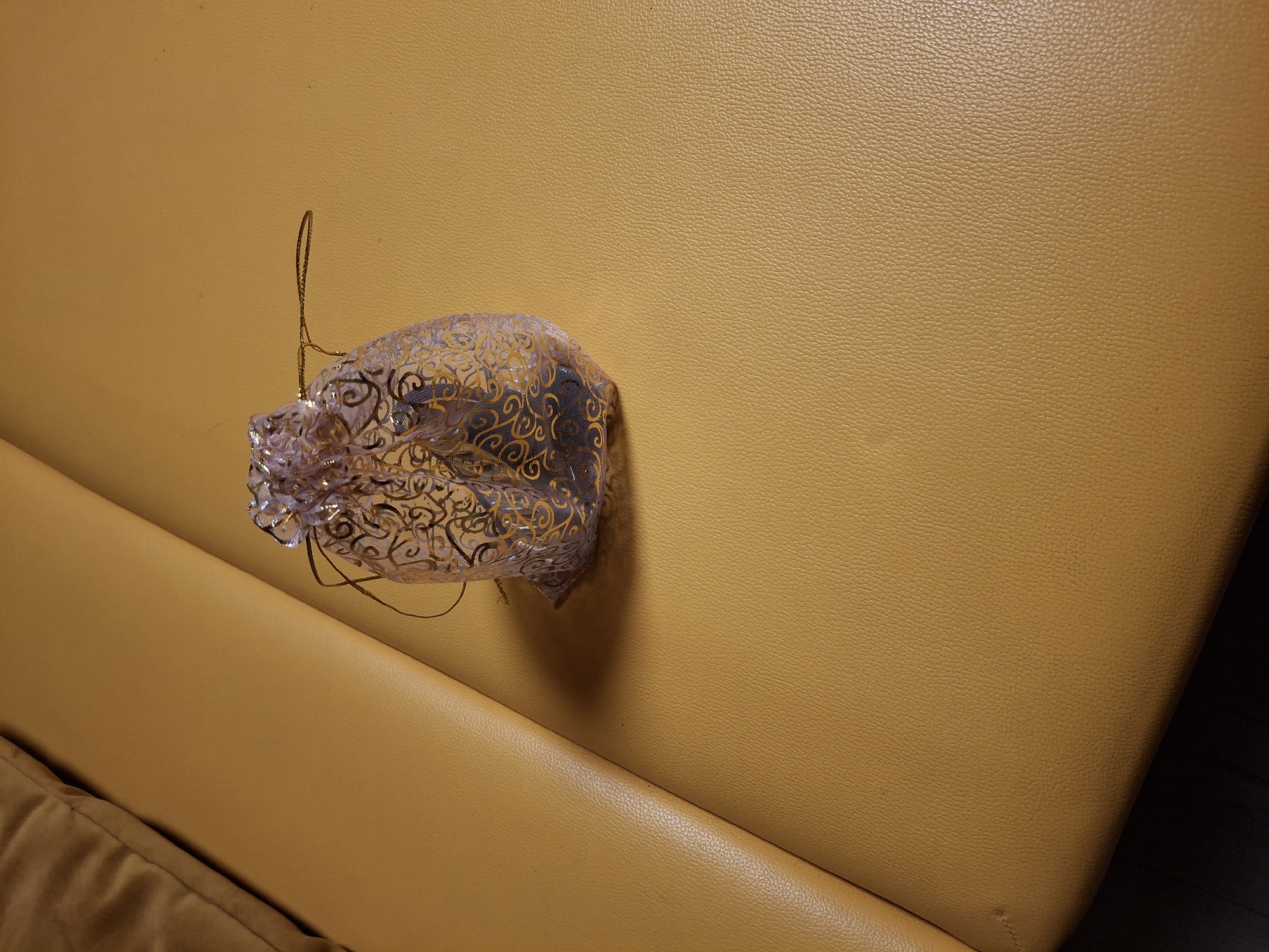 A close-up, eye-level shot shows a clear plastic ornament with a decorative gold pattern. The ornament has a string attached, suggesting it's a hanging decoration. It rests on a textured, mustard-yellow surface that appears to be upholstered, possibly a couch or chair. The surface has visible stitching in the lower left corner. The lighting casts a subtle shadow behind the ornament, indicating a light source from the upper right. The background is a smooth continuation of the textured surface, with no other objects or people visible. The image appears to be taken indoors, with no indication of time of day or weather conditions. The overall mood is neutral and focused on the object and its immediate surroundings.