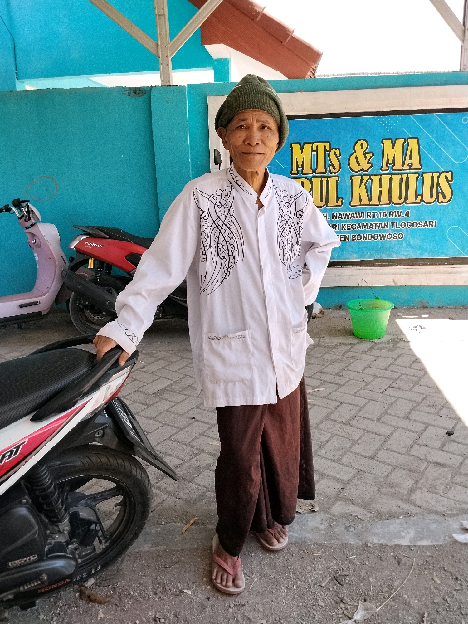 An elderly person wearing a green knit cap, a white shirt with black ornate designs, and brown trousers stands on a paved area in front of a blue wall. To the left of the person are two motorcycles, one pink and one red. Behind the person is a blue banner with yellow text that reads "MTs & MA NURUL KHULUS" and other Indonesian text. A green bucket is visible to the right of the banner. The ground is a mixture of paving stones and dirt. The scene appears to be outdoors, possibly in a schoolyard or public space in Panggungan, Indonesia.