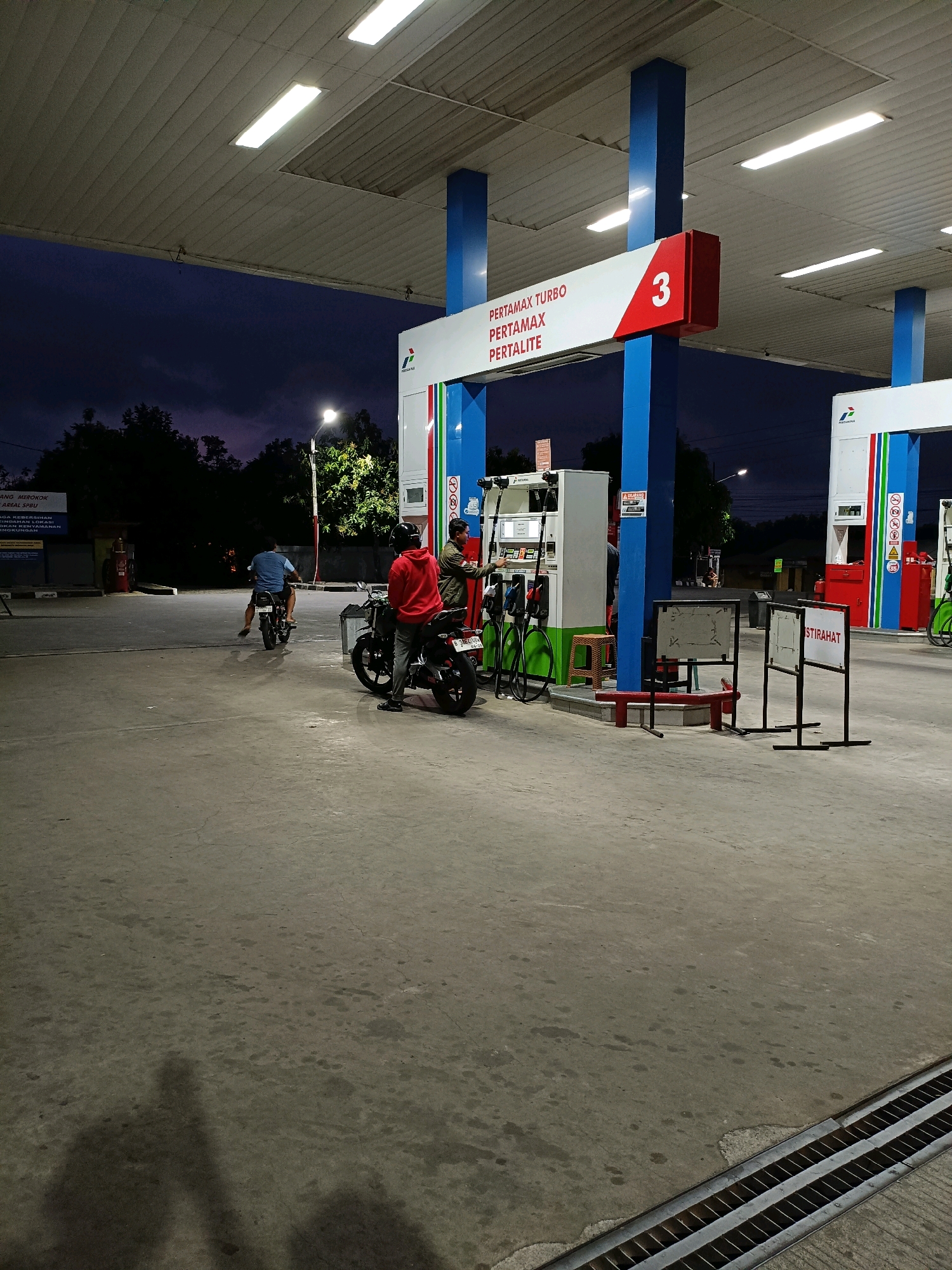 This is an image of a Pertamina gas station in Maguwoharjo, Indonesia, at dusk or night. The sky is dark blue, and the gas station is illuminated by fluorescent lights on the canopy and a streetlamp. The gas station features several fuel pumps, with one on the left and two more visible on the right, all branded Pertamina. A sign with "PERTAMAX TURBO PERTAMAX PERTALITE" and the number "3" is visible on a blue and white structure.
In the foreground, a person wearing a red hoodie is standing next to a motorcycle. To the left, another person is riding a motorcycle away from the camera. In the background, there are trees and some buildings, one of which has a sign that appears to be a "No Smoking" sign. The ground is concrete, with some oil stains visible. There are also two black metal stands with white signs, one of which reads "ISTIRAHAT". Shadows of people are visible on the ground in the lower left corner.