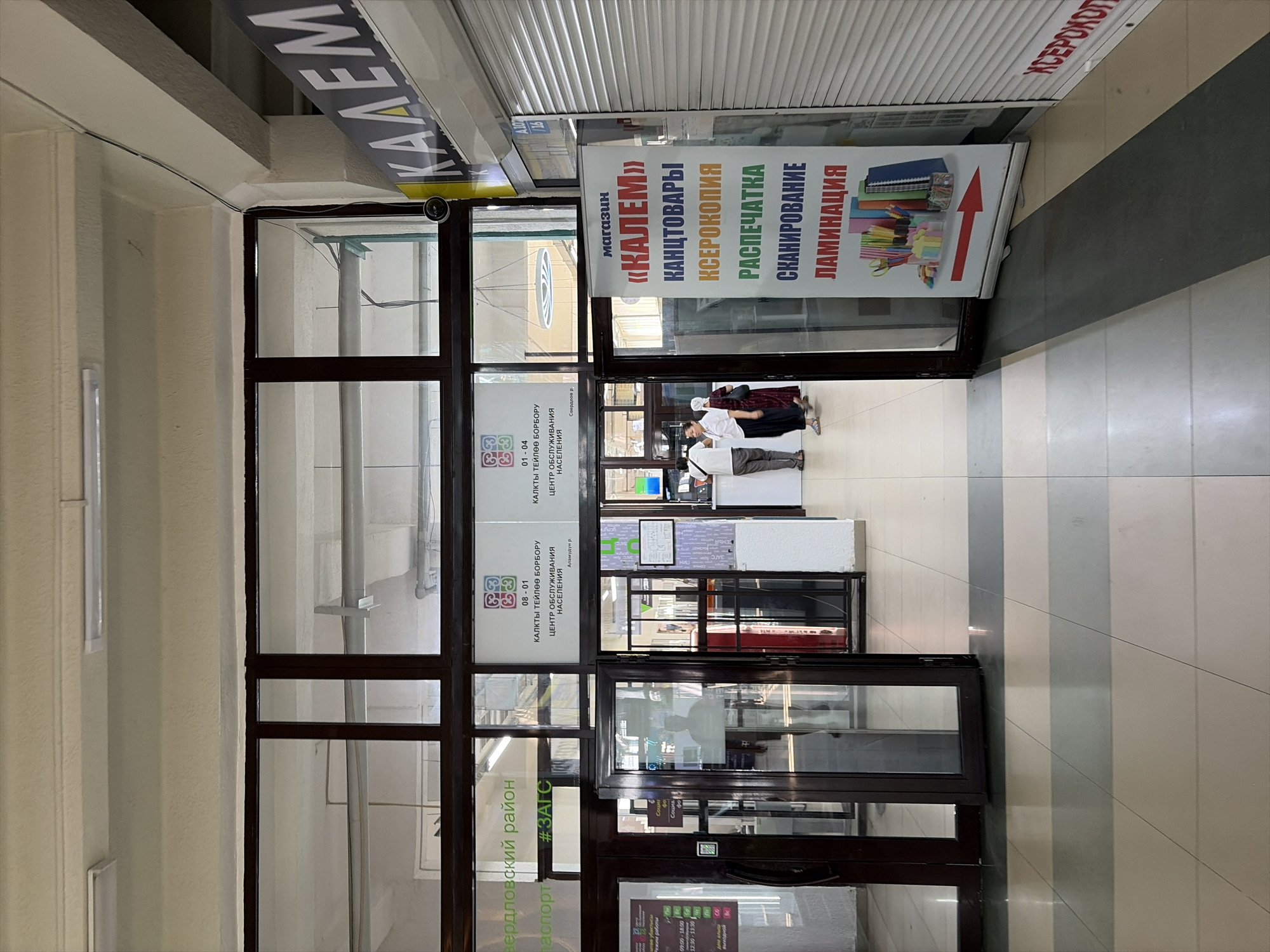 The image captures the interior of a commercial space, possibly a shopping center or market, in Bishkek, Kyrgyzstan. The perspective is from a low angle, looking down a tiled walkway towards a set of double glass doors. To the right of the entrance, a large white banner displays text in Russian, advertising "КАЛЕМ" (Kalem), a store offering stationery, photocopying, printing, scanning, and lamination services. The banner also features an illustration of stationery items and a red arrow pointing to the right. Further to the right, a closed storefront with a grey metal roller shutter is visible. Above the entrance to the left, there are signs with Cyrillic text, one of which mentions "Свердловский район" (Sverdlovsky district) and "паспорт" (passport), suggesting a government service office. Two QR codes are also visible on signs above the entrance, likely for digital information. Inside the building, through the glass doors, several people are visible, including two individuals standing near a counter. The flooring is made of large square tiles in shades of beige, grey, and dark green, arranged in a striped pattern. The lighting appears to be a mix of overhead fluorescent lights and possibly natural light filtering in from larger windows. A security camera is mounted on the upper right corner of the frame.