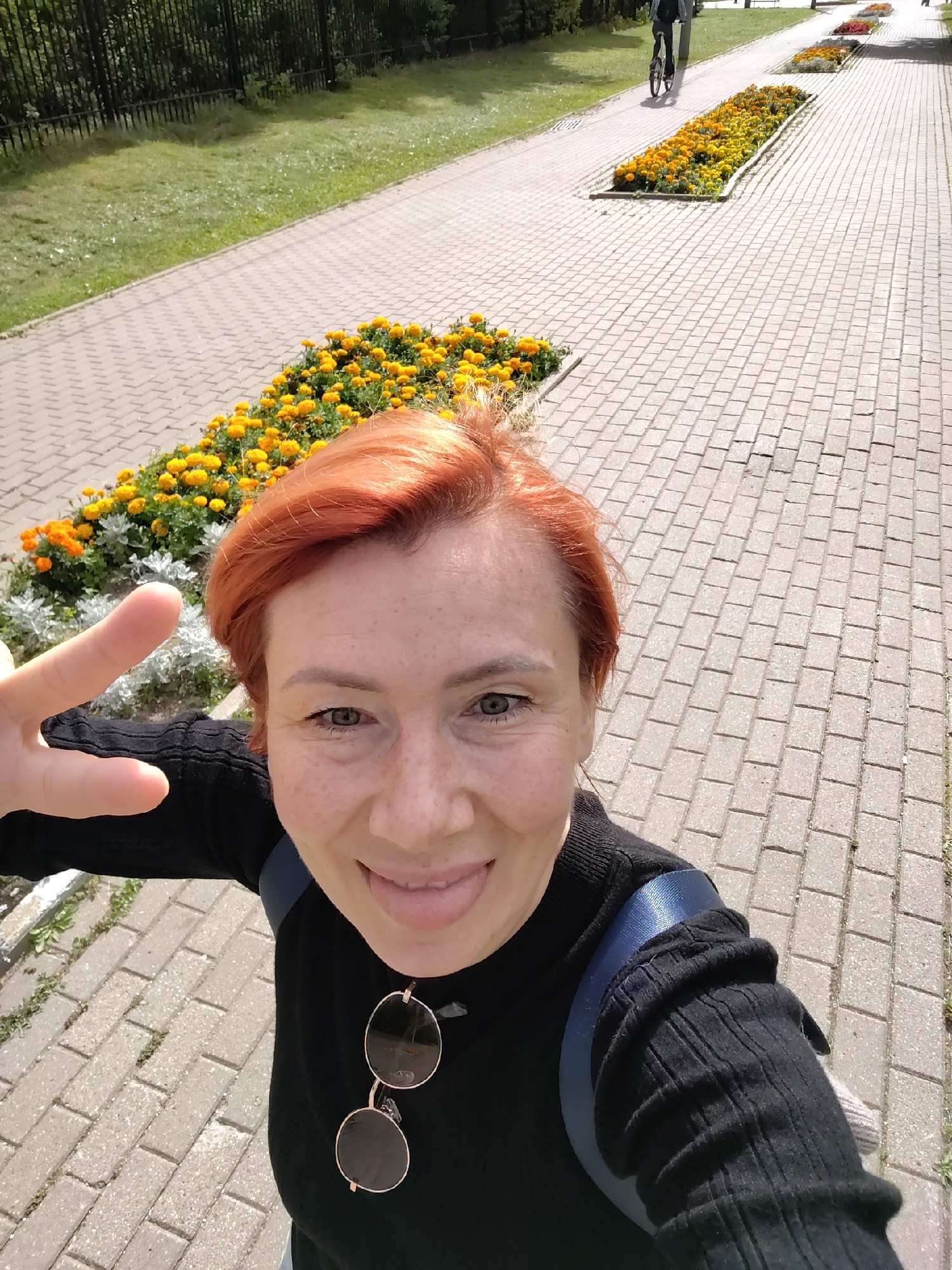 A woman with short, red hair and freckles takes a selfie outdoors. She is wearing a black long-sleeved shirt and has her tongue sticking out. A pair of round, gold-rimmed sunglasses hangs from her shirt. She is holding up two fingers in a peace sign. The background shows a paved walkway lined with flower beds of yellow and orange flowers. In the distance, a person rides a bicycle on the walkway. The location appears to be a park or public space in Zelenograd, Russia.