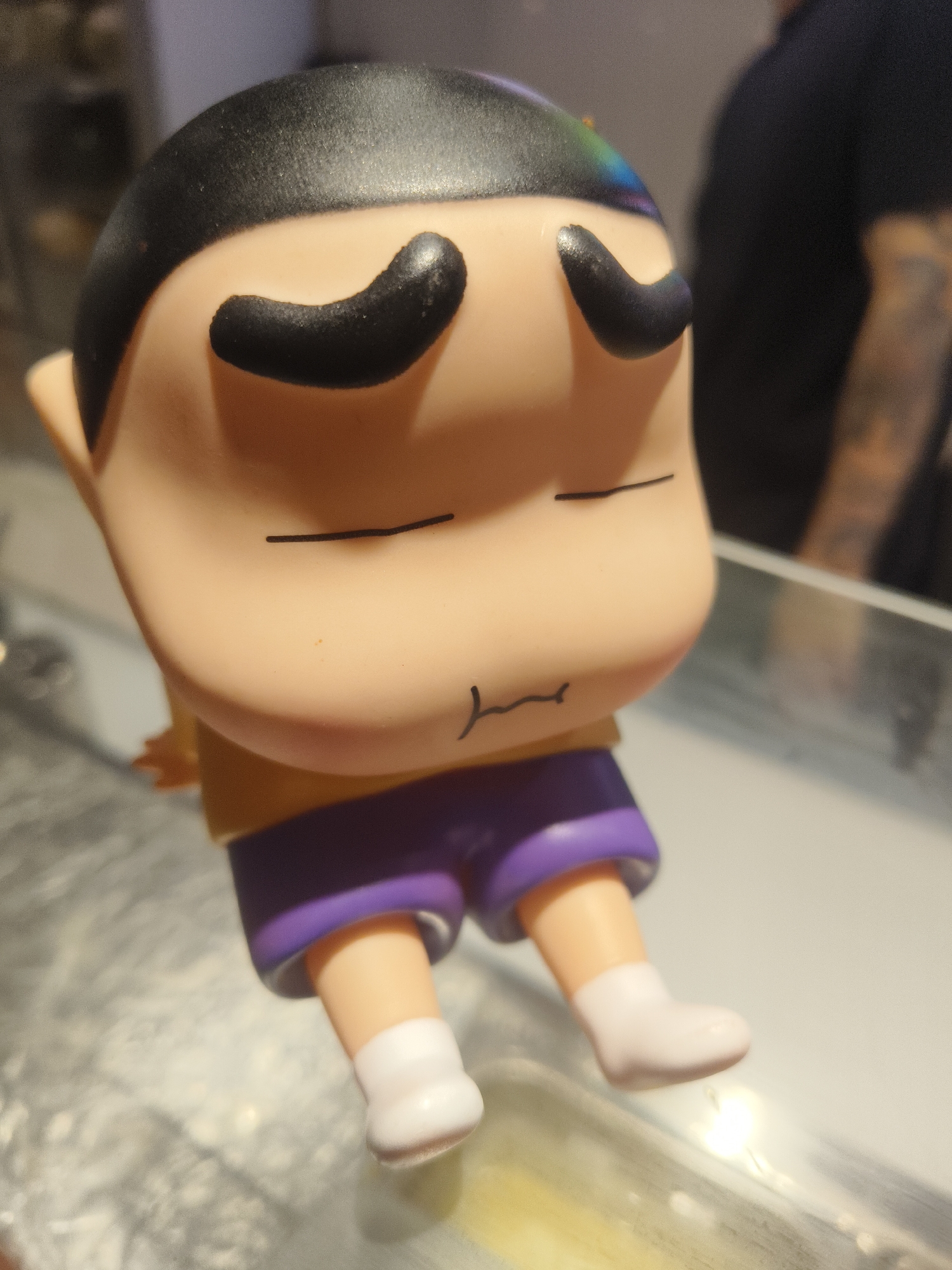 The media file shows a close-up of a small plastic figurine of a cartoon character. The figurine has a tan face with closed, dark arched eyebrows, a thin black line representing its mouth, and a black helmet-like hairstyle with a faint rainbow sheen on the top. It is wearing a yellow shirt and purple shorts, with white socks and light pink shoes. The figurine is leaning back, with its legs slightly bent, suggesting it is sitting or reclining. The background is out of focus, but appears to be an indoor setting with muted lighting. A person wearing a dark shirt is visible in the background on the right side. The overall impression is that of a playful or whimsical object, likely a toy or collectible. No visible text is present in the image.