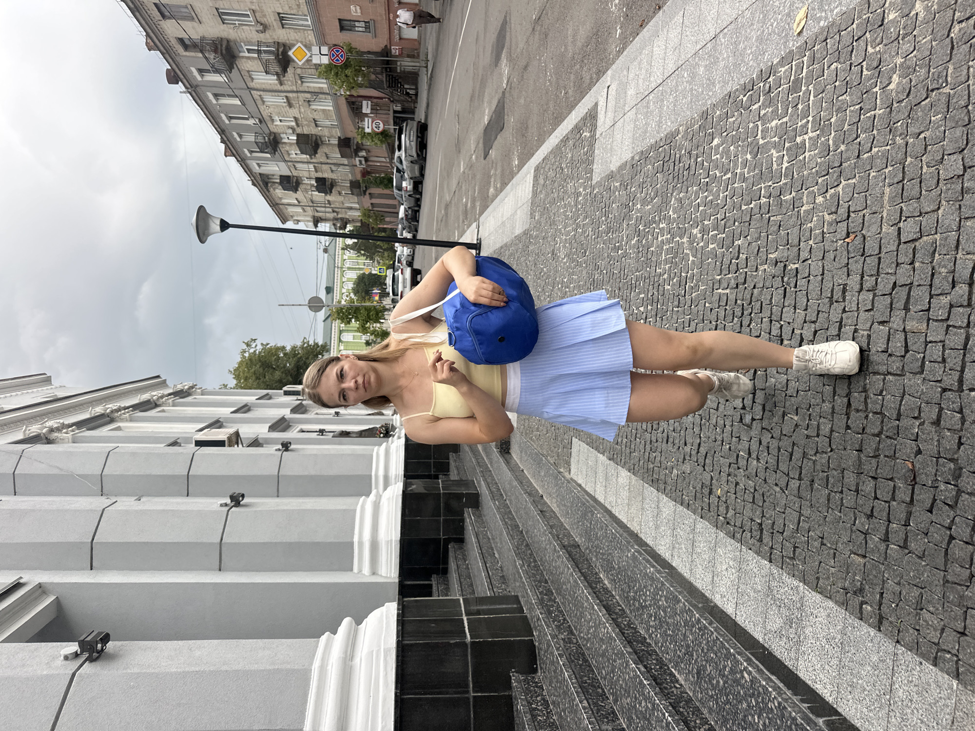 A young Caucasian woman walks down a cobblestone sidewalk in front of a building with large pillars. She is wearing a yellow tank top, a light blue pleated skirt, and white sneakers. She is carrying a large, bright blue duffel bag over her right shoulder. The building behind her has large gray pillars and a grand staircase. To her right, a street runs parallel to the sidewalk with parked cars and buildings on the opposite side. The sky is overcast. The location appears to be the Ternopil National Economic University campus in Ternopil, Ukraine, based on the architectural style of the building and its presence on a street with city infrastructure.