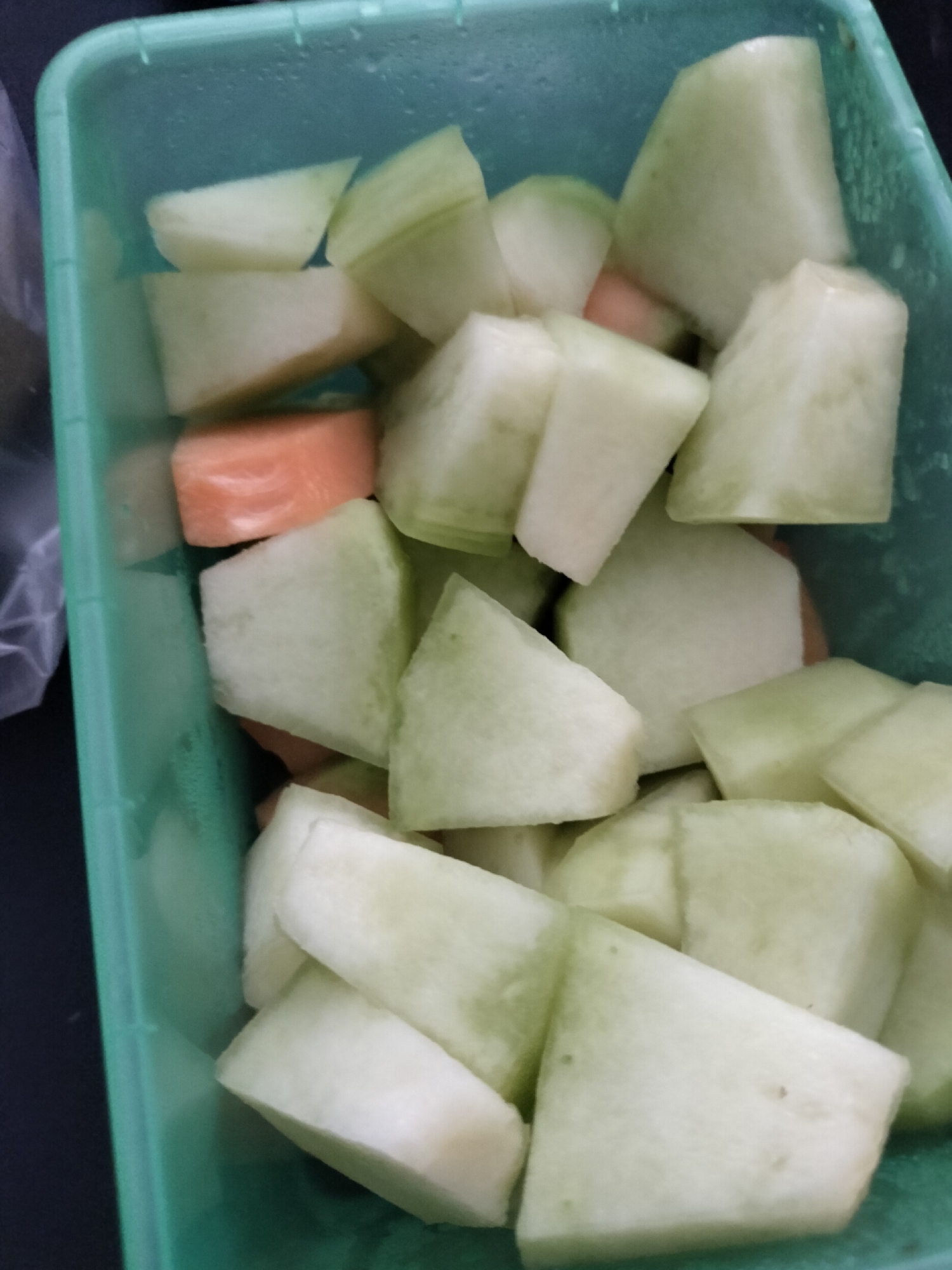 In Rantopanjang, Indonesia, a close-up shot reveals a bright green, rectangular plastic container filled with chopped honeydew melon and a few pieces of carrot. The melon pieces are irregular in shape, with some appearing to be wedges and others more cubed. The rind of the melon is visible on some pieces, showing a light green hue, while the flesh is a pale green. A few bright orange carrot pieces are interspersed among the melon. The container itself has a textured surface and appears to be slightly damp, with visible water droplets on the interior. The lighting suggests an indoor setting, possibly a kitchen or storage area.