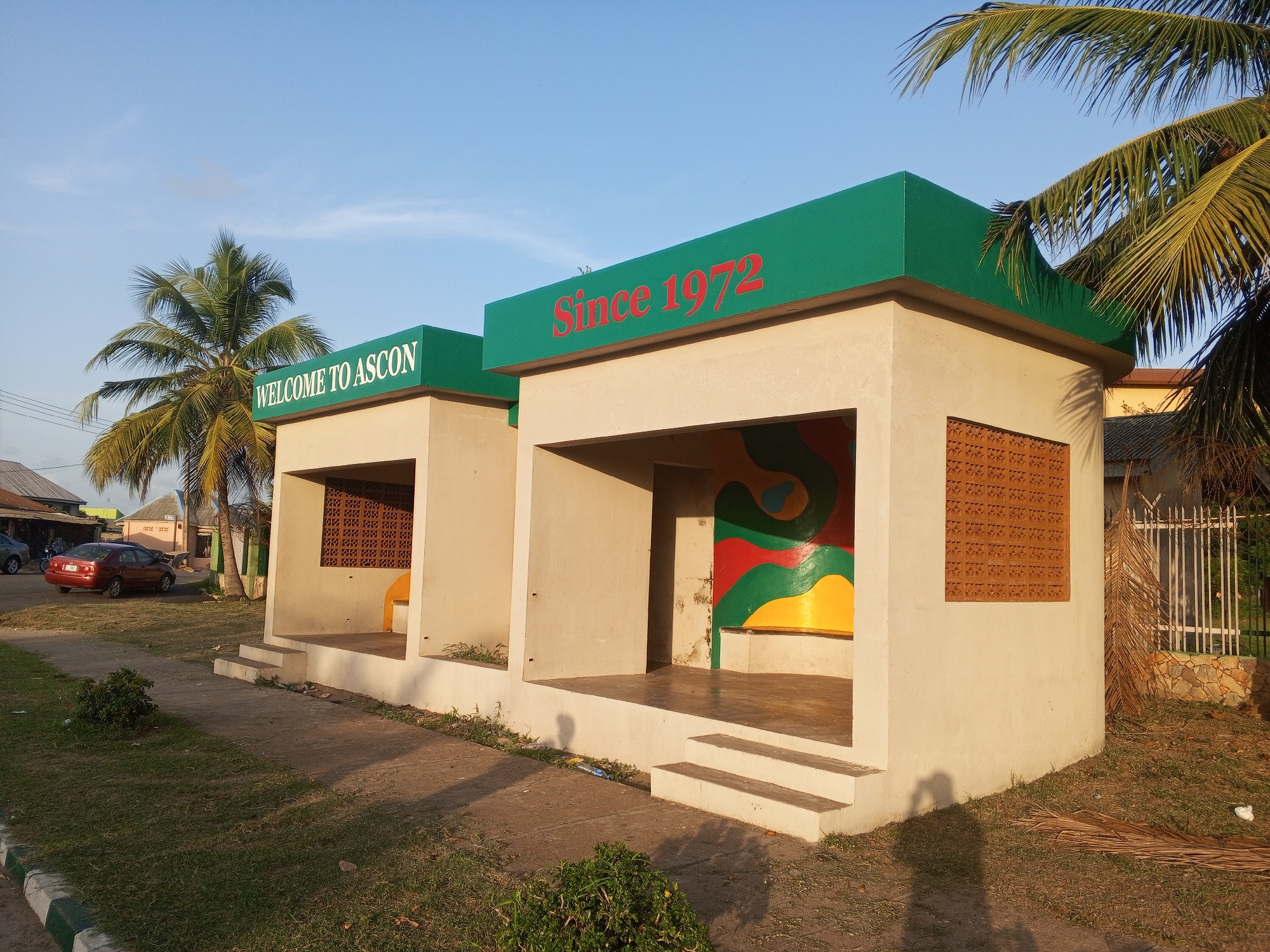 Two concrete bus stops stand side-by-side, each with a flat, green roof. The one on the left reads "WELCOME TO ASCON" in white letters on the green roof. The one on the right reads "Since 1972" in red letters. Both structures are open in the front, with tan-colored walls and steps leading up to the platform. The left bus stop has a single yellow seat. The right bus stop features a mural with green, red, and yellow colors. A decorative, patterned brick window is on the right side of each bus stop. Palm trees frame the scene. In the background on the left, there are parked cars and a building. On the right, there is a white fence and a house partially visible. The foreground consists of grass and a sidewalk. The scene is located in Tawpo, Nigeria.