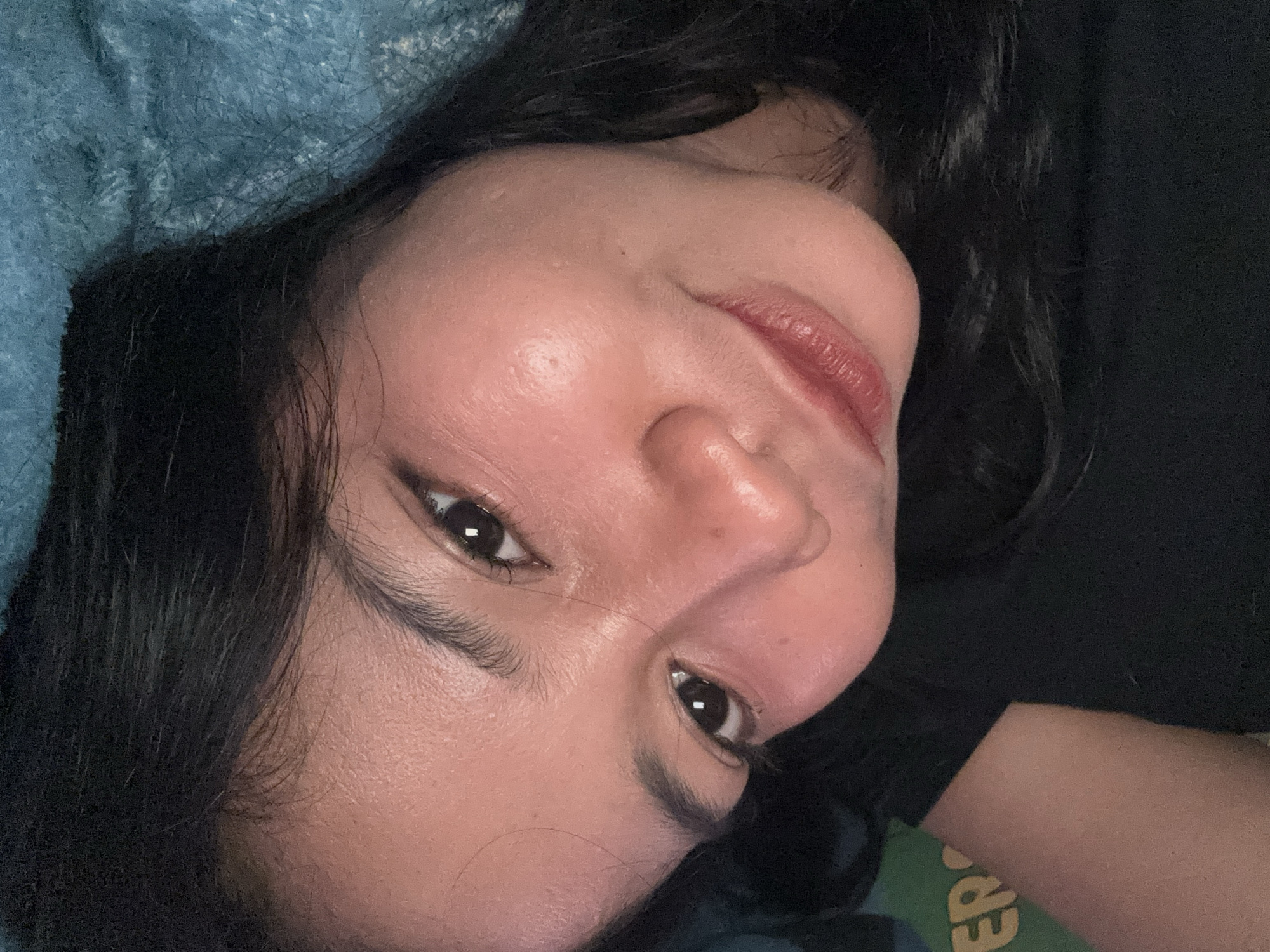A close-up, rotated portrait captures the face of a person, likely a young adult female, looking directly at the camera. Her head is tilted, giving the image an intimate and relaxed feel. She has clear, luminous skin, dark, expressive eyes with prominent eyelashes, and well-defined dark eyebrows. Her lips are a soft reddish-pink, slightly parted in a subtle, gentle smile. Fine facial hair is visible, and a small mole is present on her forehead.
Her dark, wavy hair frames her face, appearing thick and healthy. On the left side of the frame, a textured, blue fabric, resembling a blanket or cushion, is present. To the right and below her face, a dark fabric, possibly a shirt, can be seen. The background is predominantly dark, which emphasizes the subject's face.
The lighting is soft and direct, creating a gentle glow on her skin and highlighting her features. The overall mood conveyed is calm and serene. In the bottom right corner of the image, a green surface with partially visible yellow text, starting with "EP", can be seen. The full word is not discernible. The scene appears to be an indoor setting in North Jakarta, Indonesia.