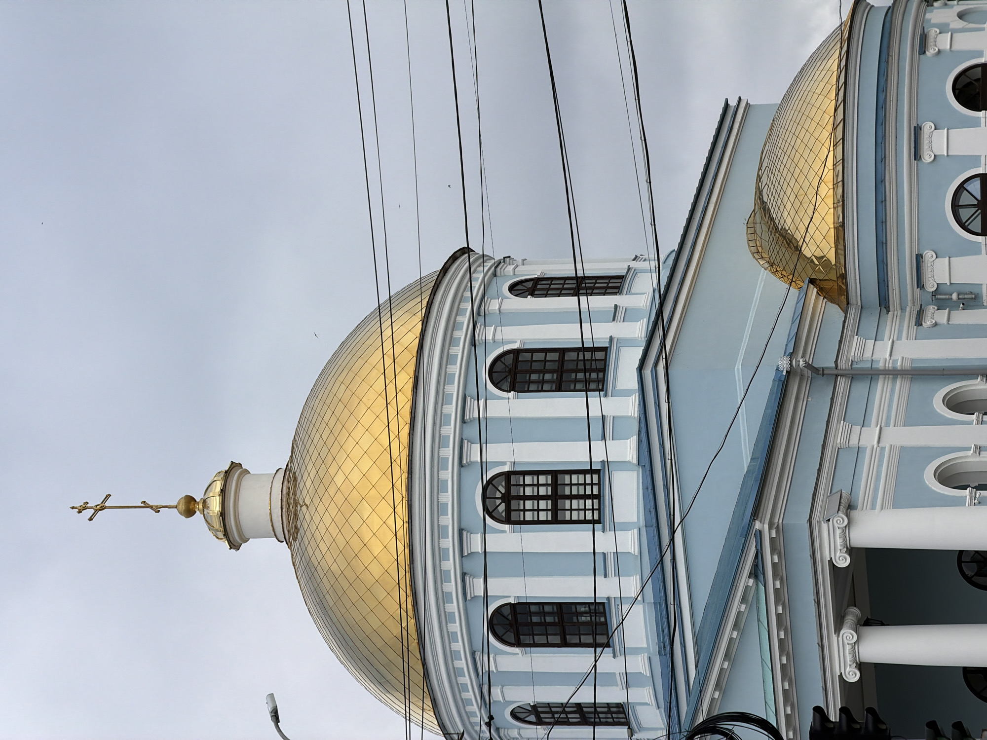 The image shows the Votkinsk Cathedral of the Holy Trinity in Votkinsk, Russia. The cathedral is a pale blue building with white architectural details. It features a large golden dome topped with a cross, and a smaller golden dome on a side section. There are arched windows with dark frames visible. The sky is overcast. Overhead, several power lines are visible, crisscrossing the frame. A traffic light and a lamppost are partially visible on the left side of the image.