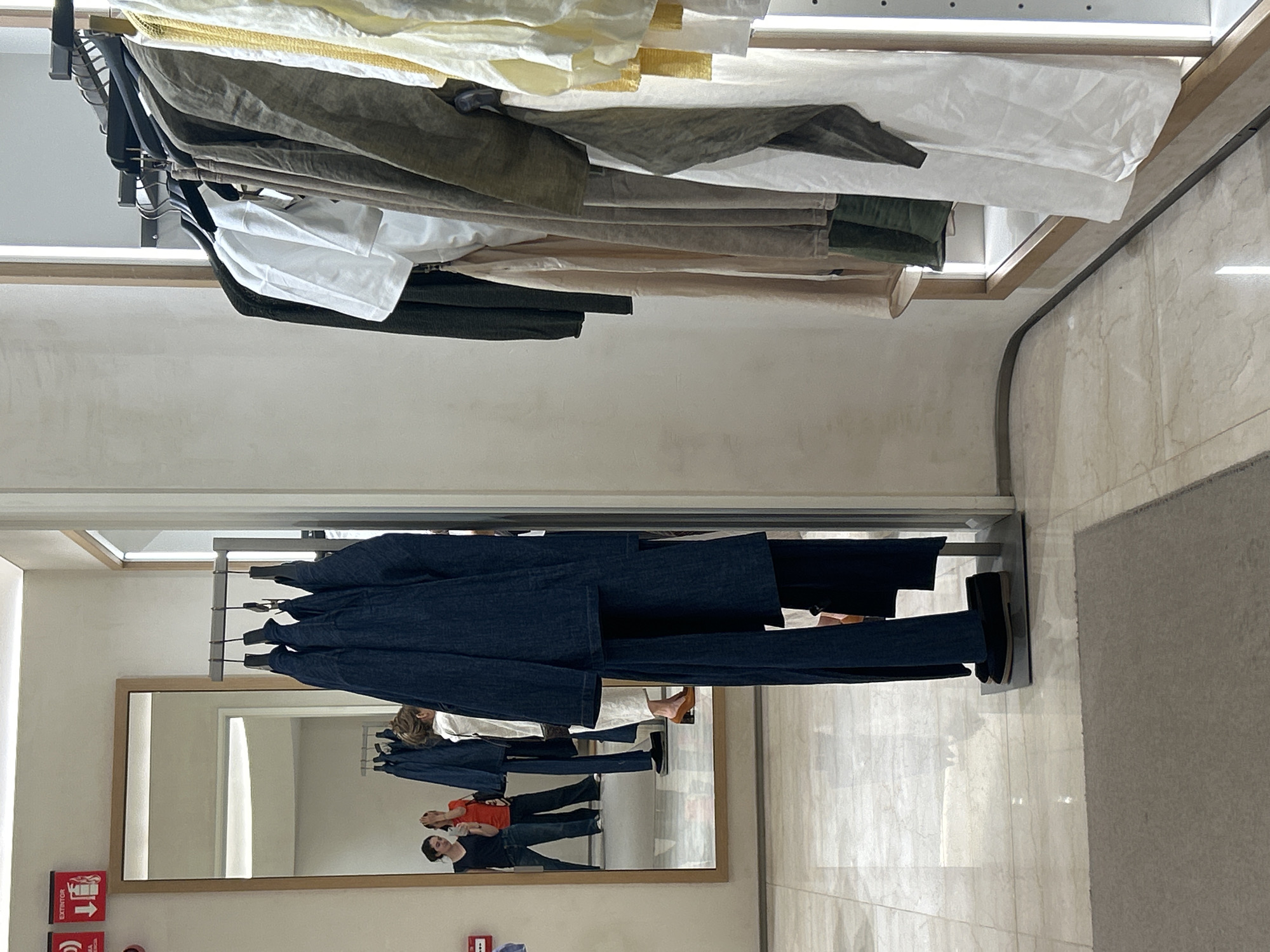 Here is a description of the image:
The image shows clothing racks in what appears to be a retail environment, possibly a clothing store. There are two racks visible, one above the other. The top rack contains light-colored garments, primarily in shades of white, cream, and light green. The bottom rack holds darker clothing, largely in blue.
In the background, a large mirror is visible, reflecting the store environment. Reflected in the mirror are at least two people; one wearing an orange shirt and the other wearing a black shirt.
The setting includes tiled flooring and a neutral-colored wall. There are signs on the wall indicating emergency exits, or extinguishers.
The image is indoors with artificial lighting, likely taken in a retail store in Unknown, Unknown.