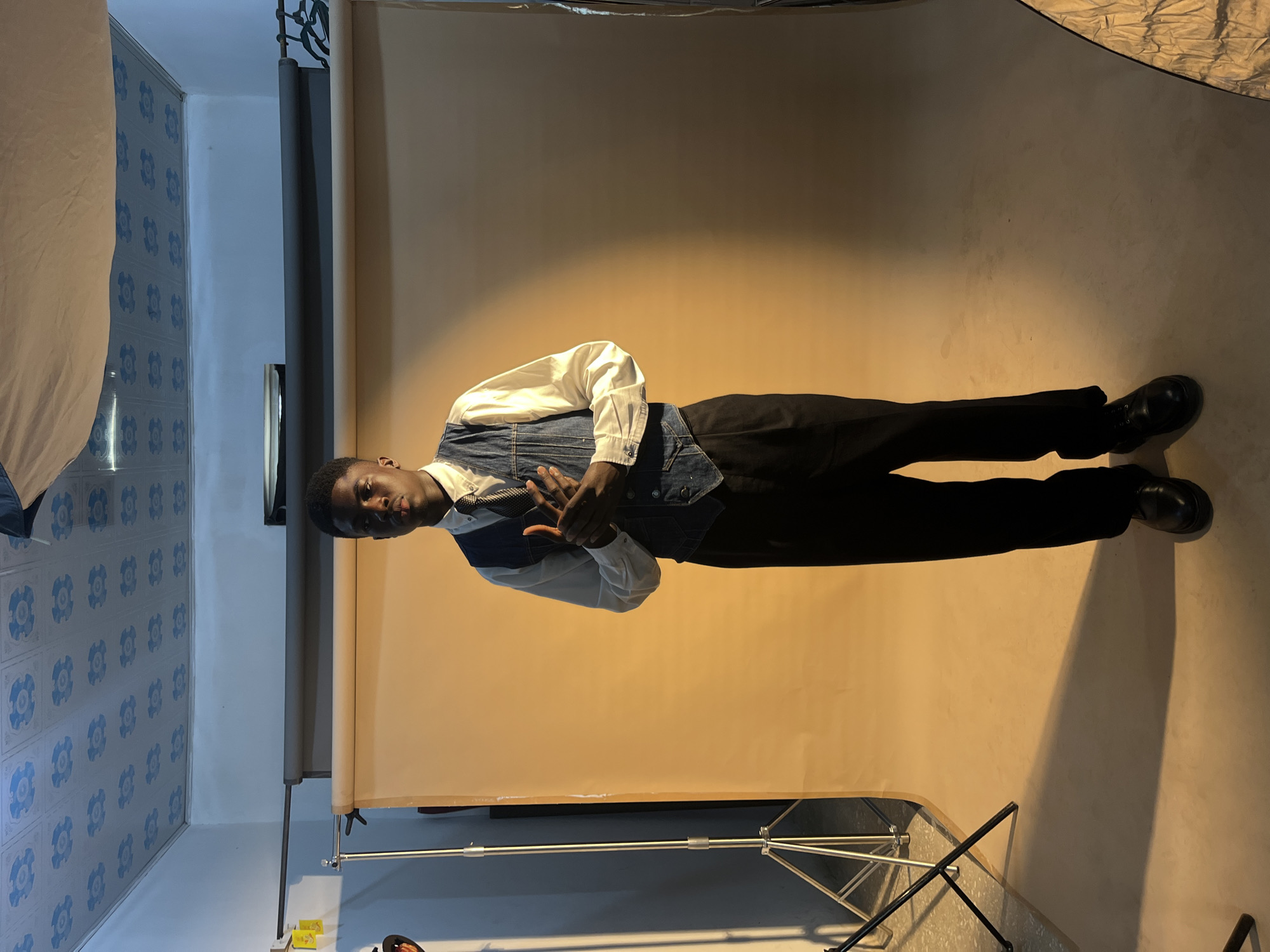 A young man is the focus of a portrait shot in what appears to be a photo studio. The individual is dressed in a white long-sleeved shirt, a denim vest, black pants, and black dress shoes. He is standing with his hands folded in front of him.
The background consists of a solid, light-tan backdrop that is likely part of a portable photography setup. Elements of studio equipment, such as the stands and lighting umbrellas, are visible around the edges of the frame. The floor is a pale, neutral color, reflecting the light from the studio lighting.
In the periphery, hints of the room outside the studio area can be seen. There is patterned blue wallpaper. This area is likely in the city of Gura Top, Nigeria.