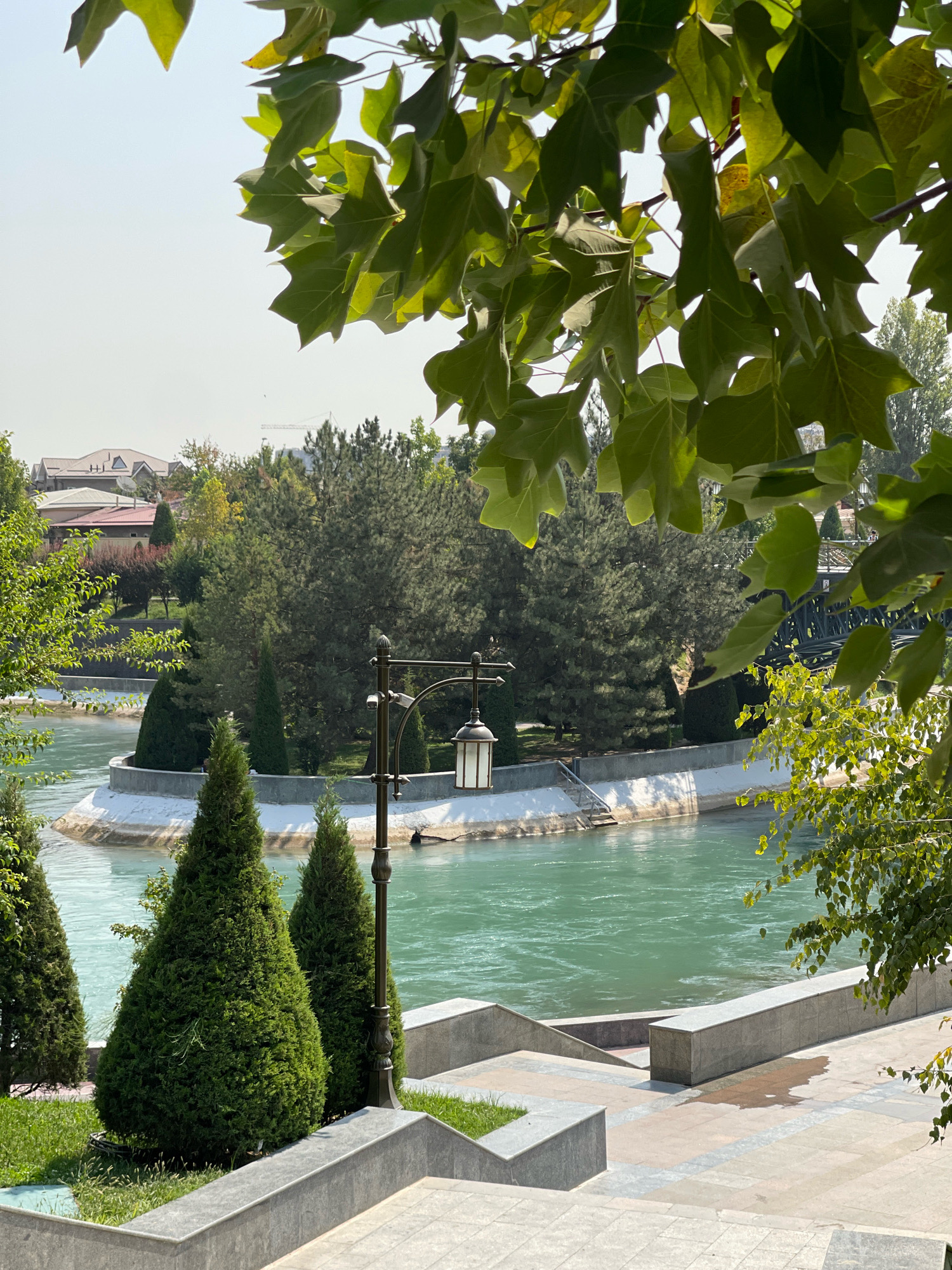 The image depicts a scene at the Ankhor Canal in Tashkent, Uzbekistan, on a clear, sunny day. The foreground features a paved area with steps and landscaping, including manicured conical evergreen trees. A classic street lamp with a lantern is visible on the right side of the paved area. Beyond the paved area, the tranquil, turquoise waters of the Ankhor Canal flow. On the opposite bank, a circular island is adorned with greenery and topped with more trees. In the background, a lush green park area extends, with mature trees and some buildings, including a residential structure with a red roof, visible in the distance. A bridge, likely part of the park's infrastructure, is partially obscured by foliage on the right side of the frame. The sky is bright and clear, indicating a warm day. There are no people visible in the image.