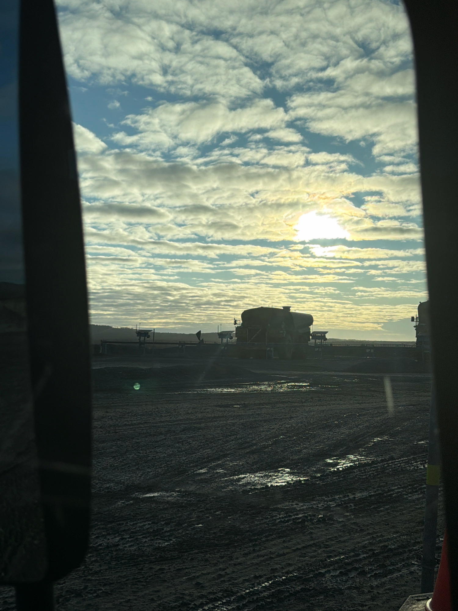 The image captures a view through a window of what appears to be a vehicle or structure, looking out onto a muddy industrial landscape under a cloudy sky. The sun is partially obscured by clouds, casting a bright light and creating a hazy effect. In the mid-ground, silhouetted industrial structures are visible, including large tanks or vessels. The ground is uneven and appears to be dirt and mud, with tire tracks evident, suggesting vehicle activity. The overall impression is one of an outdoor industrial site, possibly under construction or in operation, with overcast weather conditions. The provided information suggests the location is Gällivare, Sweden, a city known for its mining industry, which aligns with the industrial nature of the scene.