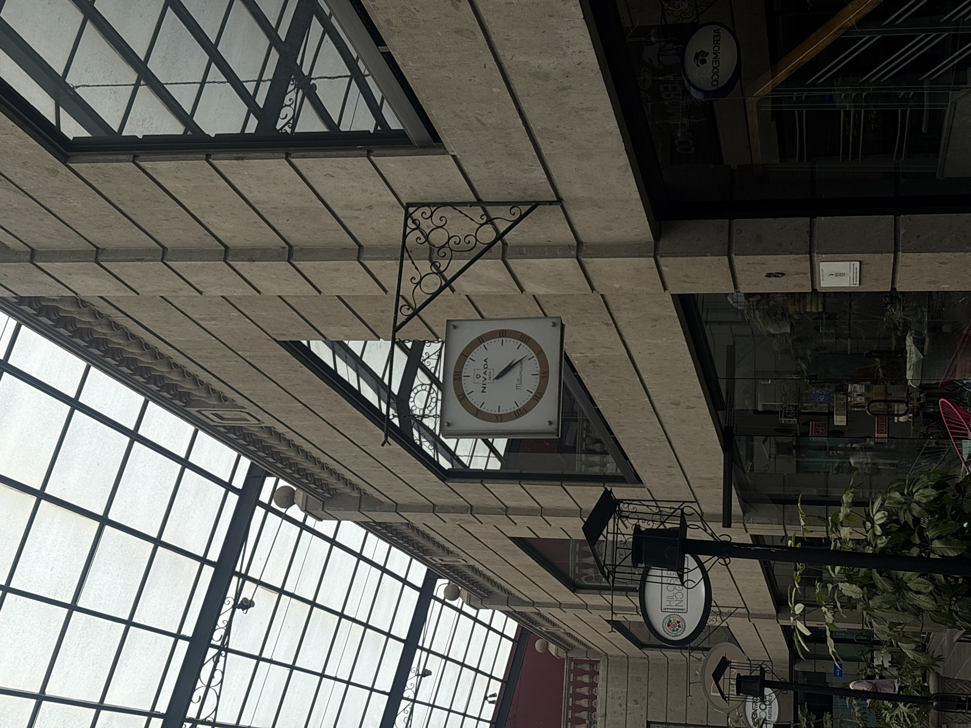 An upward-facing view captures the architectural details of an indoor or covered outdoor space, possibly a shopping arcade or plaza, in Mexico City. The dominant features are the ceiling and a suspended clock.
The ceiling consists of light-colored, rectangular stone-like panels arranged in a staggered pattern, creating a textured surface. Above and to the left, a large, expansive glass roof with a dark grid pattern allows natural light to illuminate the space. Similar glass structures are visible on the right, suggesting a continuous overhead covering.
A prominent square white clock, adorned with Roman numerals, hangs from the ceiling. Its hour hand points towards X (10) and the minute hand towards II (2), indicating approximately 10:10. The clock face displays "NIVADA" and "Millionaire" in black text. It is supported by an ornate, scrolled black metal bracket attached to the ceiling panels.
Further down and to the right, a black metal post supports a circular sign. This sign features a colorful, geometric, circular logo and the text "HILOS DE LÚA," with "DISEÑO A MANO" and "HECHO EN MÉXICO" inscribed below it. Below this, a smaller, indistinct circular sign is also visible. Reflections in a nearby glass surface or window show reversed text, including "YEBOMEXICO."
The overall lighting suggests daytime. There are no people visible, indicating a quiet moment in the location. Various other decorative black metal elements and hints of greenery from plants add to the ambiance.