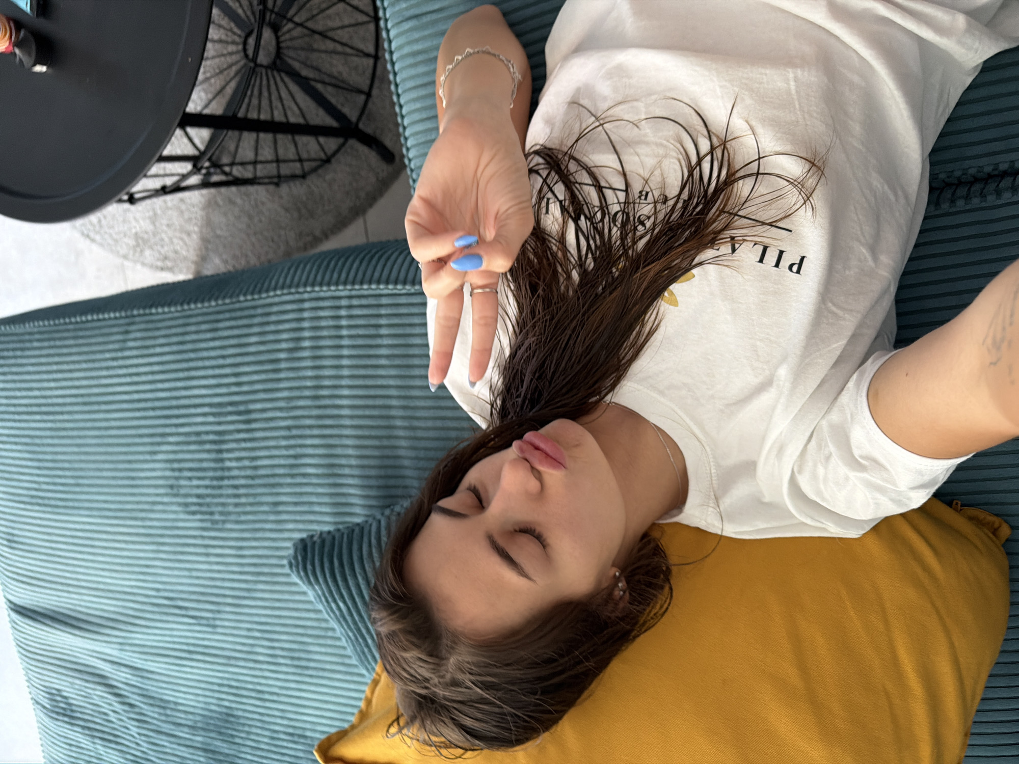 A young woman lies on a sofa with a yellow pillow under her head. She is wearing a white t-shirt with black text that is partially visible, reading "PILAT" and what appears to be a floral design. Her eyes are closed, and her lips are puckered. Her right hand is raised, displaying bright blue fingernails, with her index and middle fingers extended in a peace sign, and her thumb, ring finger, and pinky finger curled. She is wearing two rings on her left ring finger and a bracelet on her right wrist. Her dark hair is spread across the t-shirt. The sofa is teal and ribbed. In the background, a black round table with a wire base is visible, along with a fuzzy rug. The lighting suggests it is daytime.