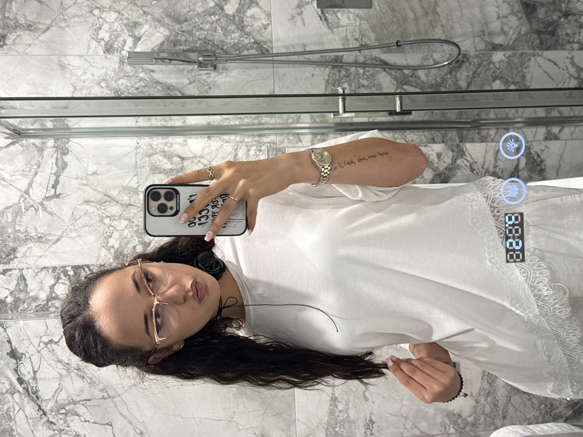 A young woman with dark hair and glasses is taking a selfie in a bathroom. She is wearing a white top with lace trim. Her left hand is raised to her hair, and her right hand is holding a phone, displaying a mirror image of herself. A watch is visible on her left wrist, and there is a tattoo on her arm. The bathroom has marble walls and a glass shower door. The scene appears to be in a private residence, not a public space within Shumen, Bulgaria.