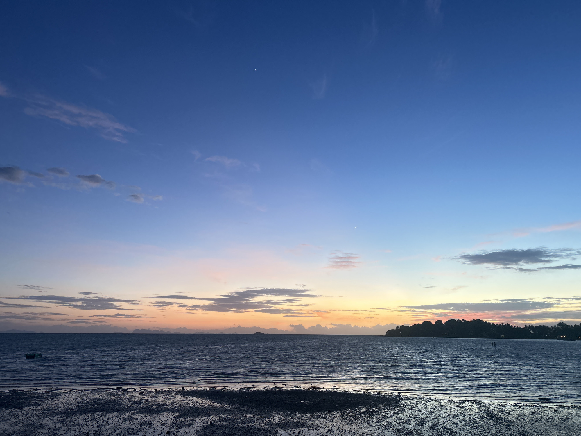 This image captures a serene sunset over the ocean in Ko Pha-ngan District, Thailand. The sky transitions from a deep blue at the top to soft hues of pink, orange, and yellow near the horizon, with scattered clouds adding texture. A faint crescent moon is visible in the upper right quadrant of the sky. The sea, a deep blue, exhibits gentle ripples, reflecting the colors of the sky. The foreground consists of a muddy, pebble-strewn shoreline. In the distance, a silhouetted landmass with trees lines the horizon, and a small island is visible further to the left. Two figures, appearing as small silhouettes, can be seen in the water on the right side of the frame. A small boat is also present on the left side of the image.