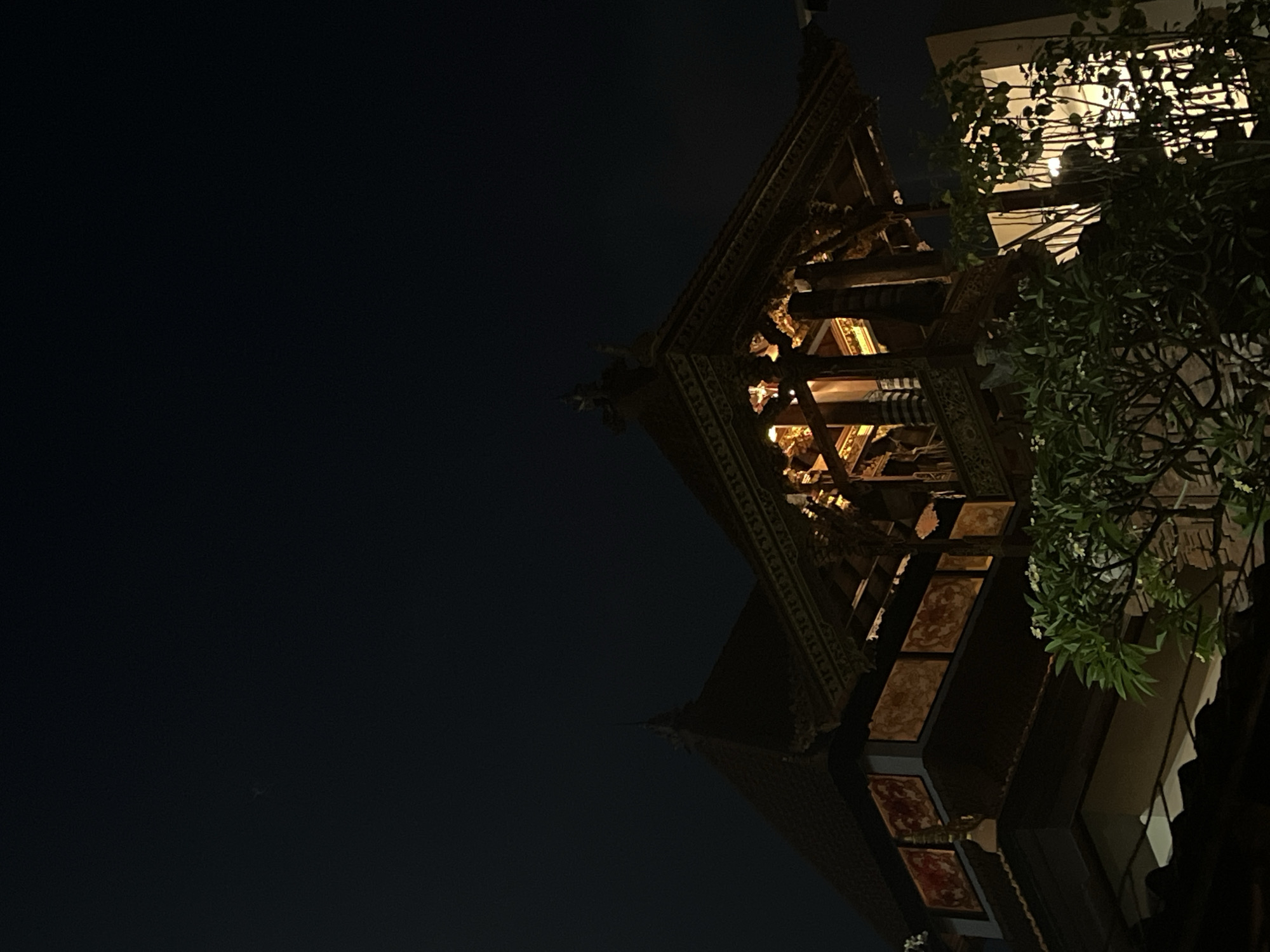 This low-angle, nighttime shot captures the upper floors of a building with traditional Indonesian architecture, possibly a temple or resort, in North Kuta, Indonesia. The structure features ornate wooden carvings and decorative panels with red and gold motifs. Warm interior lighting illuminates the details of the upper level, highlighting its intricate craftsmanship. In the foreground, lush green foliage with small white flowers partially obscures the lower parts of the building, adding a natural element to the scene. The night sky above is a deep, dark blue, with a faint speck of light, possibly a star, visible in the upper left quadrant. The overall impression is one of serene, illuminated architecture against a dark, natural backdrop.