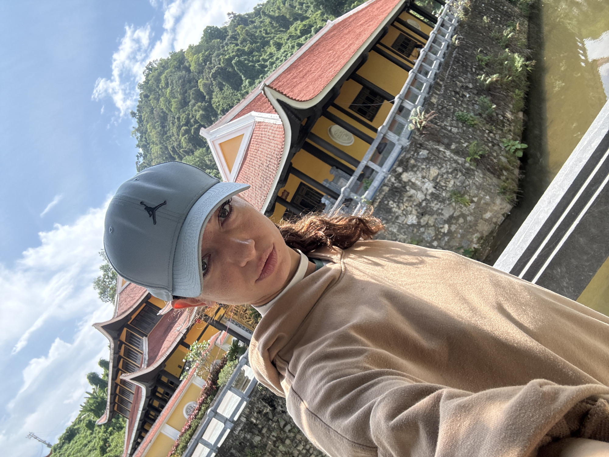 A person wearing a light gray Jordan cap and a beige sweater is positioned in the foreground, presumably taking a selfie. In the background is a traditional Vietnamese temple building, characterized by its orange tiled roof and yellow walls with black accents. The temple complex is situated next to a body of water and green foliage, appearing to be a lush, mountainous landscape. The setting is likely outdoors with a clear sky featuring some clouds. This is possibly in or around Đà Nẵng, Vietnam.