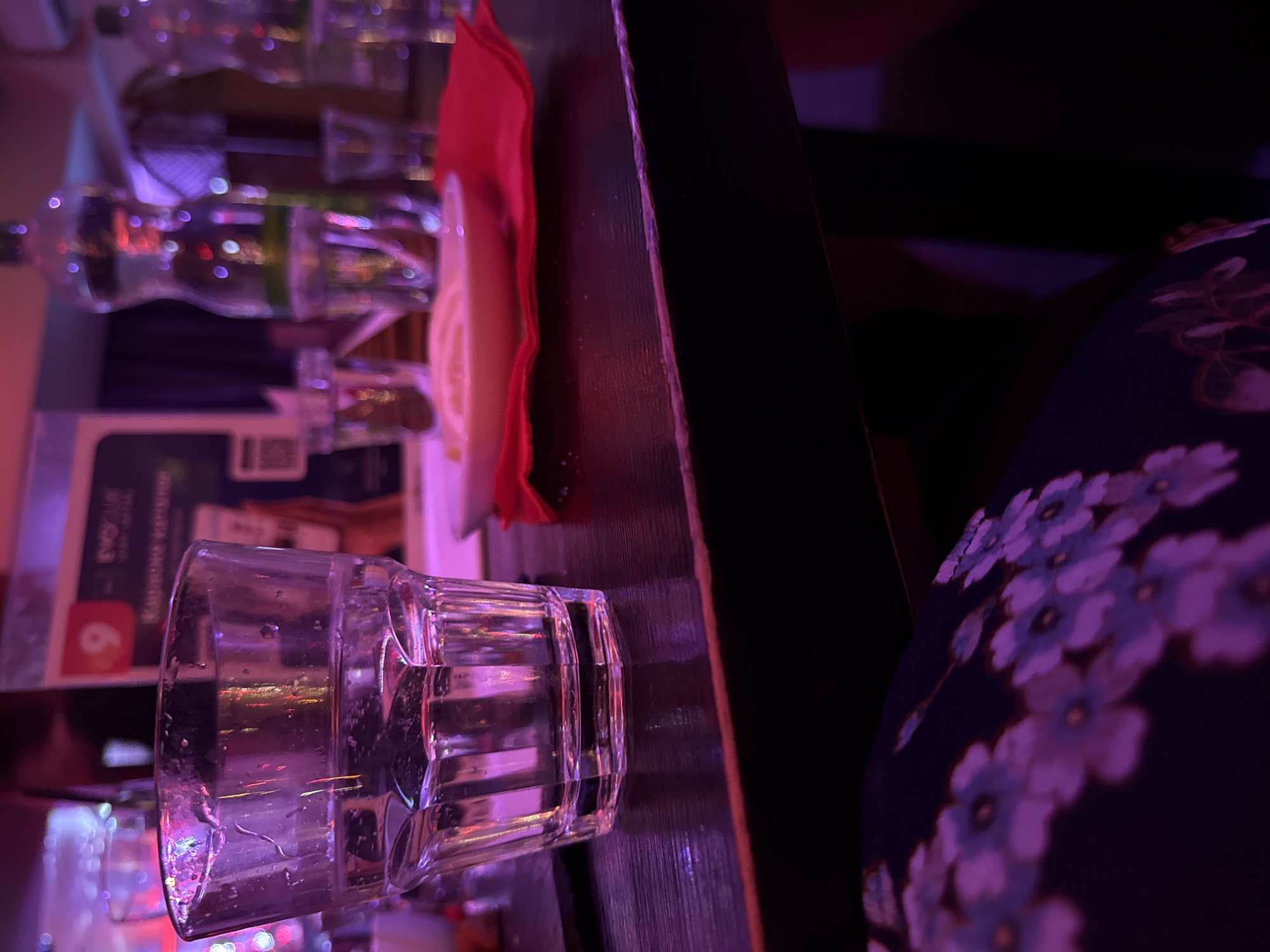 The image depicts a bar scene, possibly in Votkinsk, Russia, though this cannot be confirmed without more information. A faceted clear glass sits near the edge of a dark-stained wooden bar top, likely containing water or a clear liquid. Behind the glass are several bottles of liquor and a folded red napkin. A portion of a dark blue garment with purple and white floral patterns is visible to the right of the bar top. The lighting is dim, with a purple hue illuminating the scene.