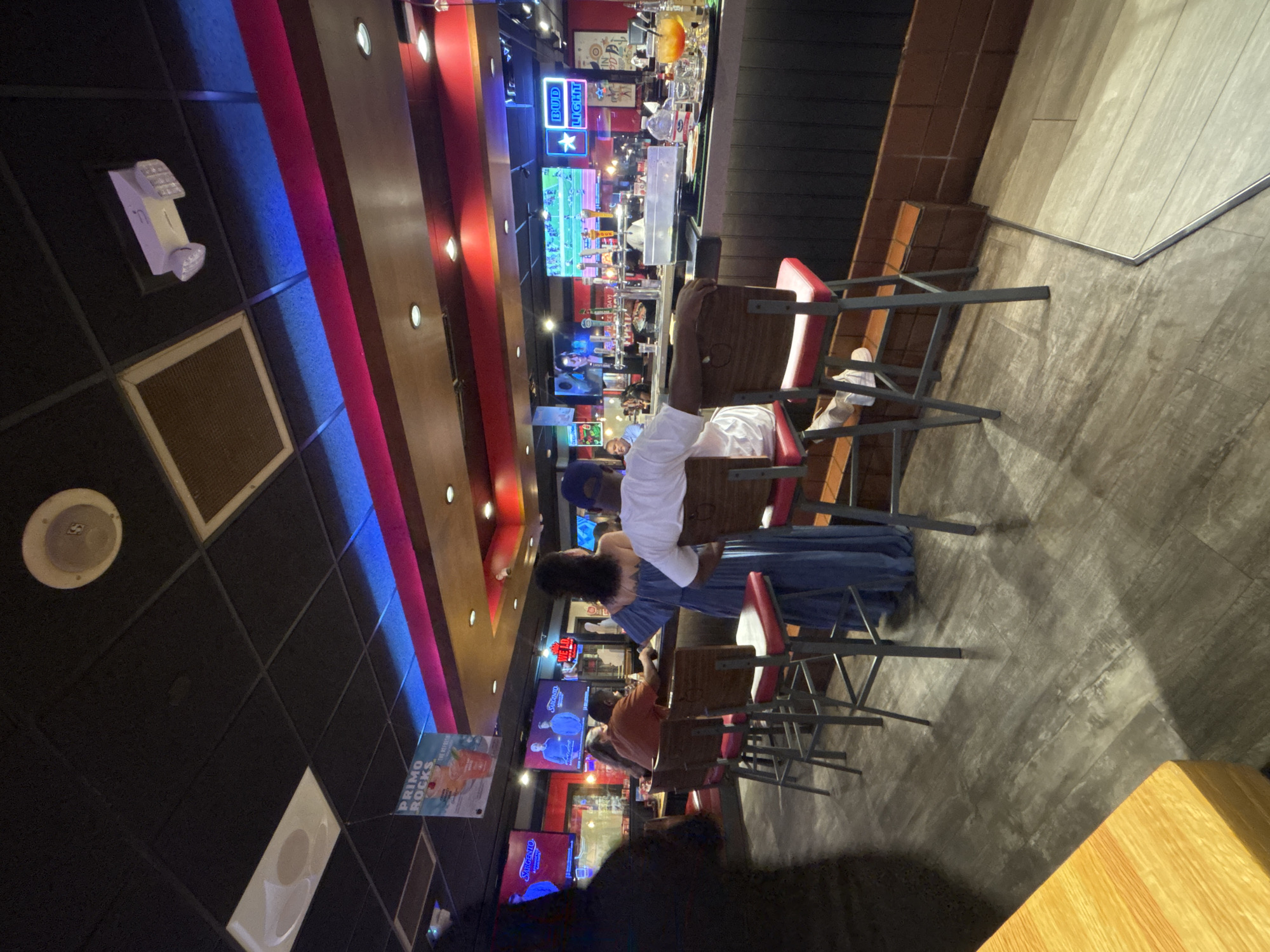 The image shows the interior of a bar or restaurant in Grand Prairie, Texas, USA. The space features a bar counter with patrons seated on red bar stools. Overhead lighting and decorative neon lights create a distinctive ambiance, with multiple television screens displaying content on the back wall behind the bar. Customers are visible at the bar and in the background, interacting with the setting. The floor has a grey finish.