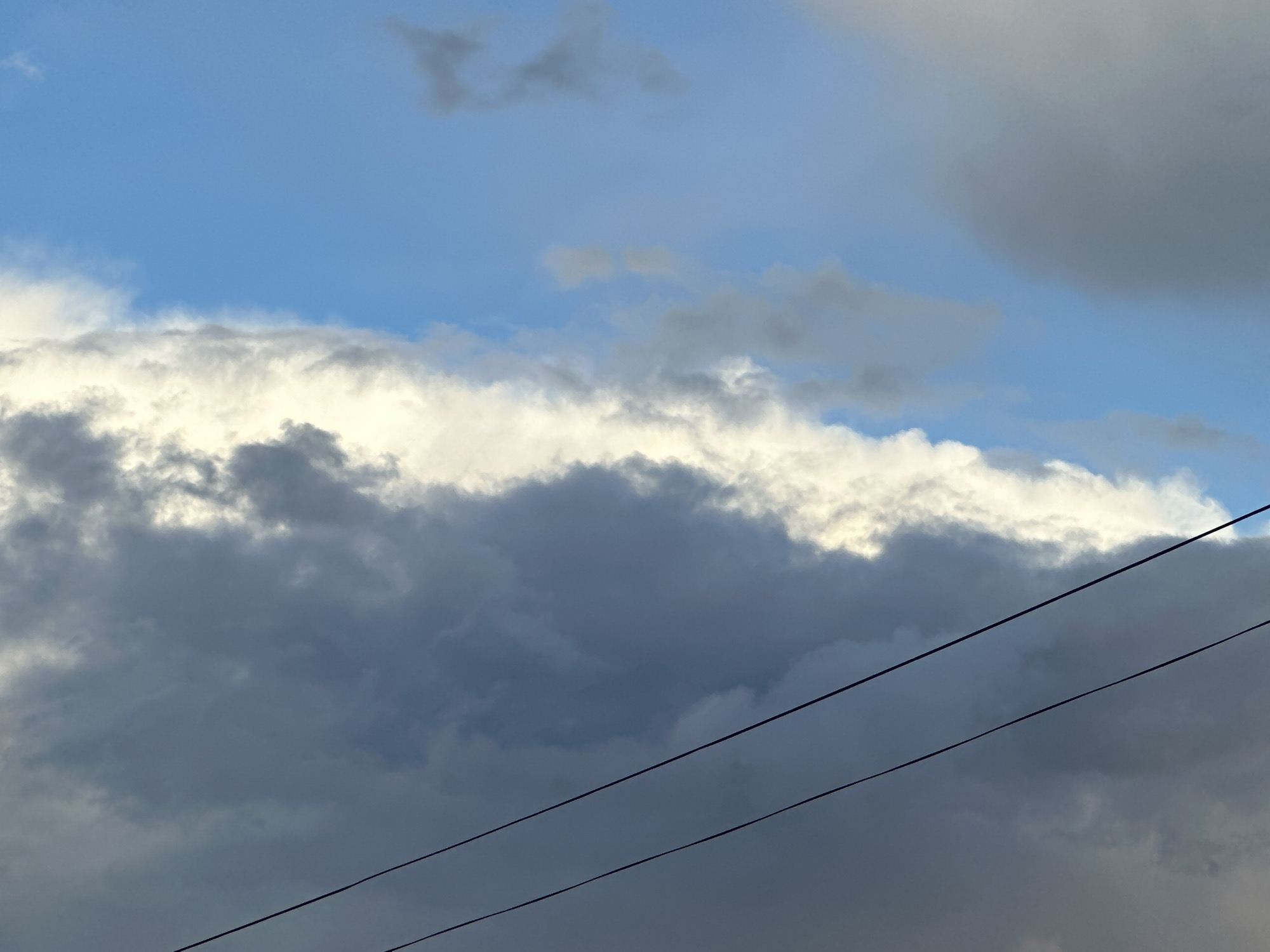 The image shows a section of sky with predominantly dark, stormy clouds. A sliver of blue sky is visible above the clouds. Two diagonal black power lines cut across the lower right portion of the frame. The location appears to be in an urban area, indicated by the presence of power lines, in the city of Unknown, Unknown.