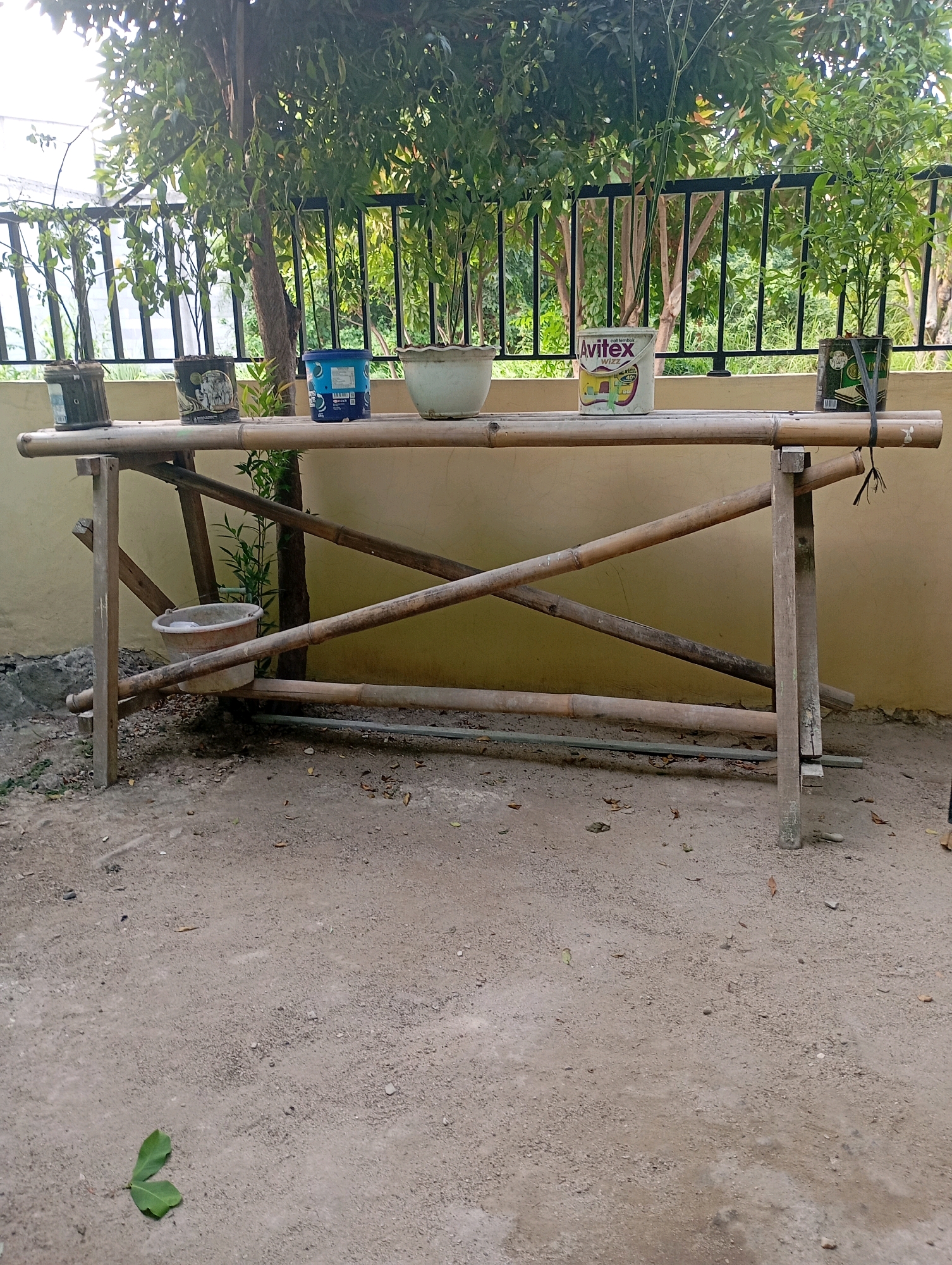 The image depicts an outdoor scene in Babelan, Indonesia, featuring a handmade bamboo table. The table is constructed from several bamboo poles, with a flat surface supported by diagonal bracing. On the table, there are four potted plants, one of which is a can with a label that reads "Avitex Wizz." A small, light-colored bucket is placed beneath the table. In the background, a beige wall is visible, with a black metal fence and lush green foliage beyond. The ground is covered in sand and dirt, with scattered leaves. The overall setting suggests a casual outdoor space, possibly a backyard or garden.