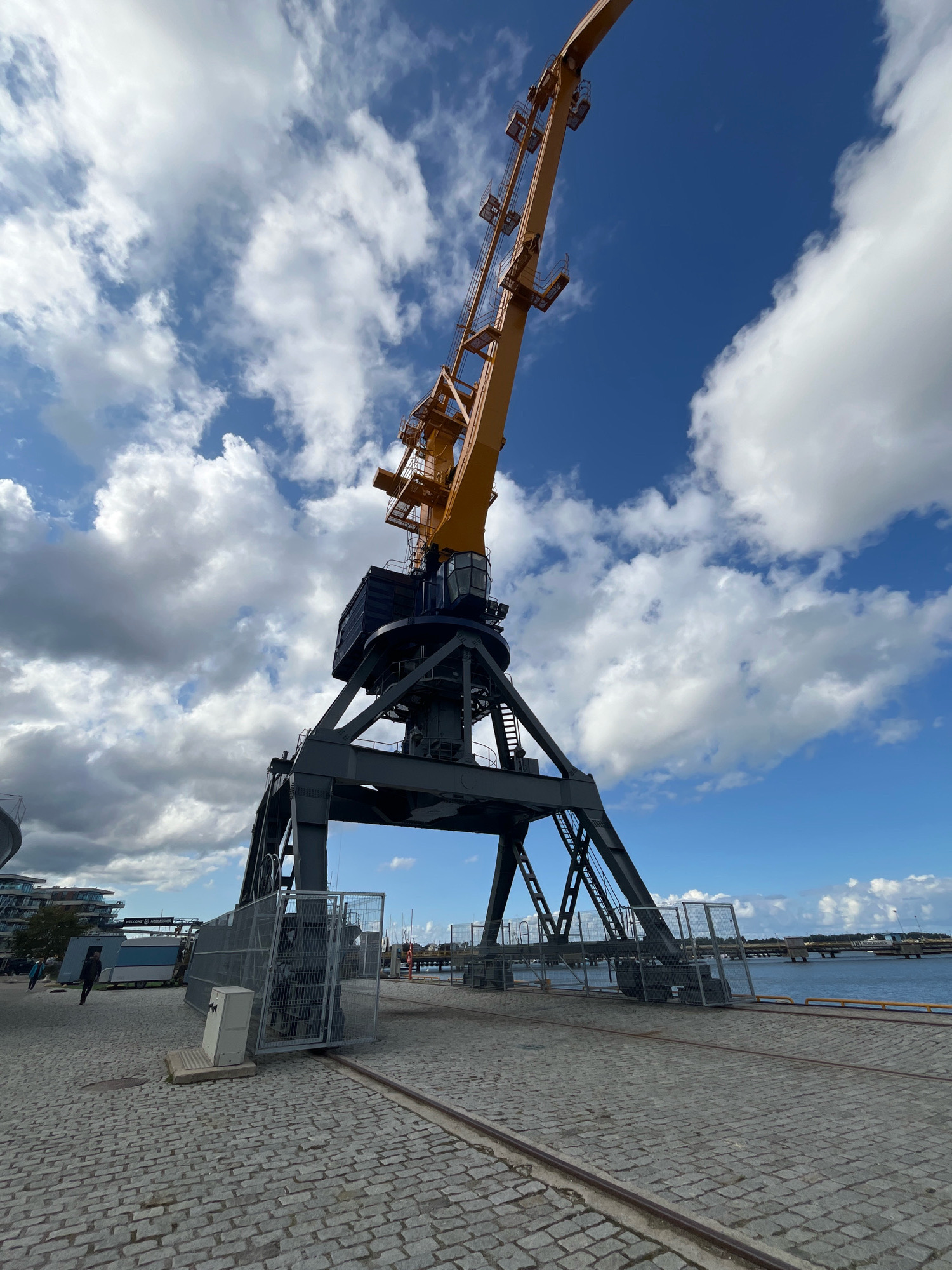 A low-angle, full shot captures a large, industrial crane in a port area. The crane's dark gray metal structure forms a prominent base, with angled legs supporting a dark blue cabin and an extended, bright yellow boom. The boom is angled upwards and to the right, against a dynamic sky filled with white, puffy clouds against a bright blue background. The ground in the foreground is paved with cobblestones and features parallel metal tracks, indicating the crane's mobility. To the left, a fenced-off area encloses equipment. In the midground, a body of water stretches out, with a bridge or pier visible in the distance. Buildings and some people are visible in the far left background, suggesting an urban or semi-urban port environment. The overall scene suggests a functional port or industrial site. This scene is located at the Tallinn Port, in Tallinn, Estonia.