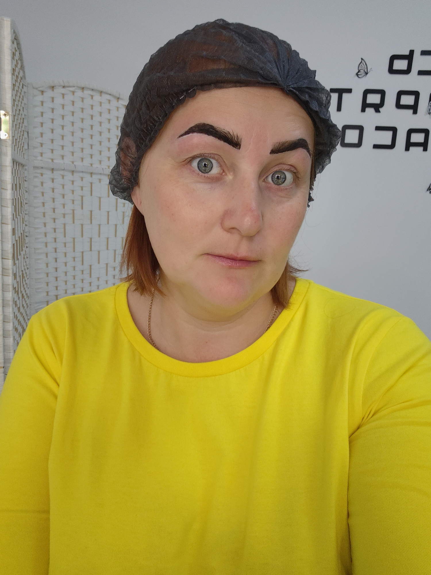 A head-and-shoulders portrait shows a woman indoors, possibly in a beauty salon in Марковский, Russia. She looks directly at the camera with wide, green eyes and a neutral, slightly surprised expression. Her reddish-brown hair is covered by a disposable black hairnet. She is wearing a bright, long-sleeved yellow t-shirt, and a thin gold chain is visible around her neck. Her eyebrows appear freshly done, possibly microbladed or tattooed, with a slight reddish outline visible around the dark, bold brows.
In the background, to the left, there is a white, wicker-patterned room divider or screen. To the right, a white wall displays partial black text in a stylized font, including "D J" at the top, and below it, "TRAQ" and "OJA". A black butterfly silhouette is visible next to "TRAQ". The lighting suggests an indoor setting, likely with artificial light.