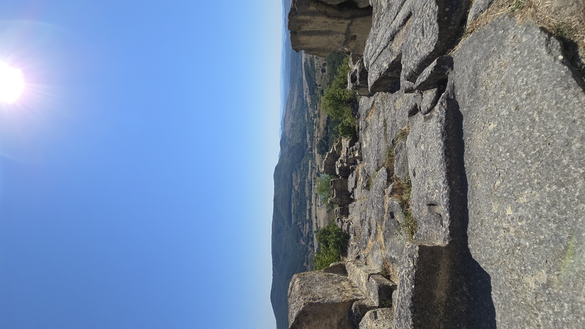 The image depicts a vast, clear blue sky with a bright sun in the upper right corner, casting a radiant glow. Below the sky, a panoramic view unfolds, showcasing a rugged, rocky foreground that appears to be part of ancient ruins. Large, weathered stone blocks are scattered across the ground, with some forming a makeshift pathway or foundation.

In the mid-ground, a verdant landscape stretches out, characterized by rolling hills covered in dense green vegetation and scattered trees. Fields of varying shades of green and brown suggest cultivated land. A body of water, possibly a lake or a wide river, is visible in the distance, reflecting the sunlight. The background features more distant, hazy mountains and hills that fade into the horizon.

The ruins in the foreground are significant, with massive stone slabs suggesting a historical site. The overall impression is one of ancient history set against a beautiful natural backdrop. The location is identified as Gorna Krepost, Bulgaria. The image appears to be taken on a sunny day, as indicated by the bright sun and clear sky. There are no people or animals visible in the scene. The image is taken from a high vantage point, offering an expansive view of the surrounding terrain.