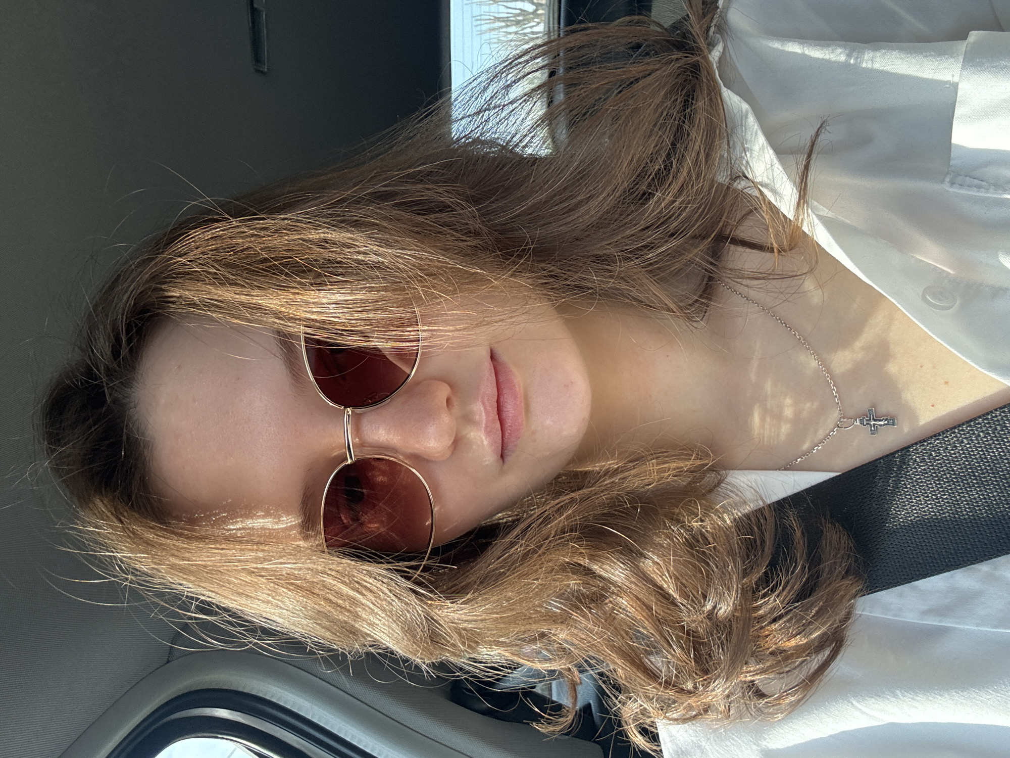 A woman wearing sunglasses and a white button-down shirt is in a car. She has brown hair and is wearing a silver necklace with a cross pendant. A black seatbelt crosses her chest. The location is likely not in Perm, Russia, as there is nothing to indicate a specific location. The scene is indoors, inside a car.