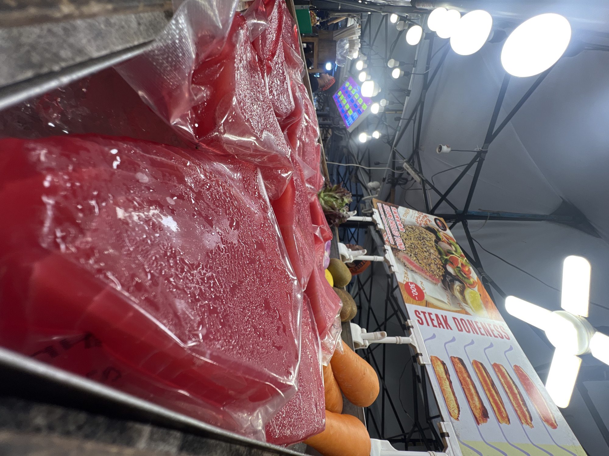 The image depicts a street food stall in Karon, Thailand, with a focus on a display of raw tuna steaks. The tuna, cut into large blocks, is individually wrapped in plastic and covered in condensation, suggesting it is kept chilled. In the background, a sign advertises "STEAK DONENESS" with images of cooked steaks, and a price of "300.-" is visible. Other produce, including carrots and a potato, are also on display. Several bright overhead lights illuminate the scene, typical of an outdoor market. No people are clearly visible in the immediate foreground, though a blurred figure can be seen in the background. The overall impression is one of a vibrant, bustling market environment specializing in fresh seafood.