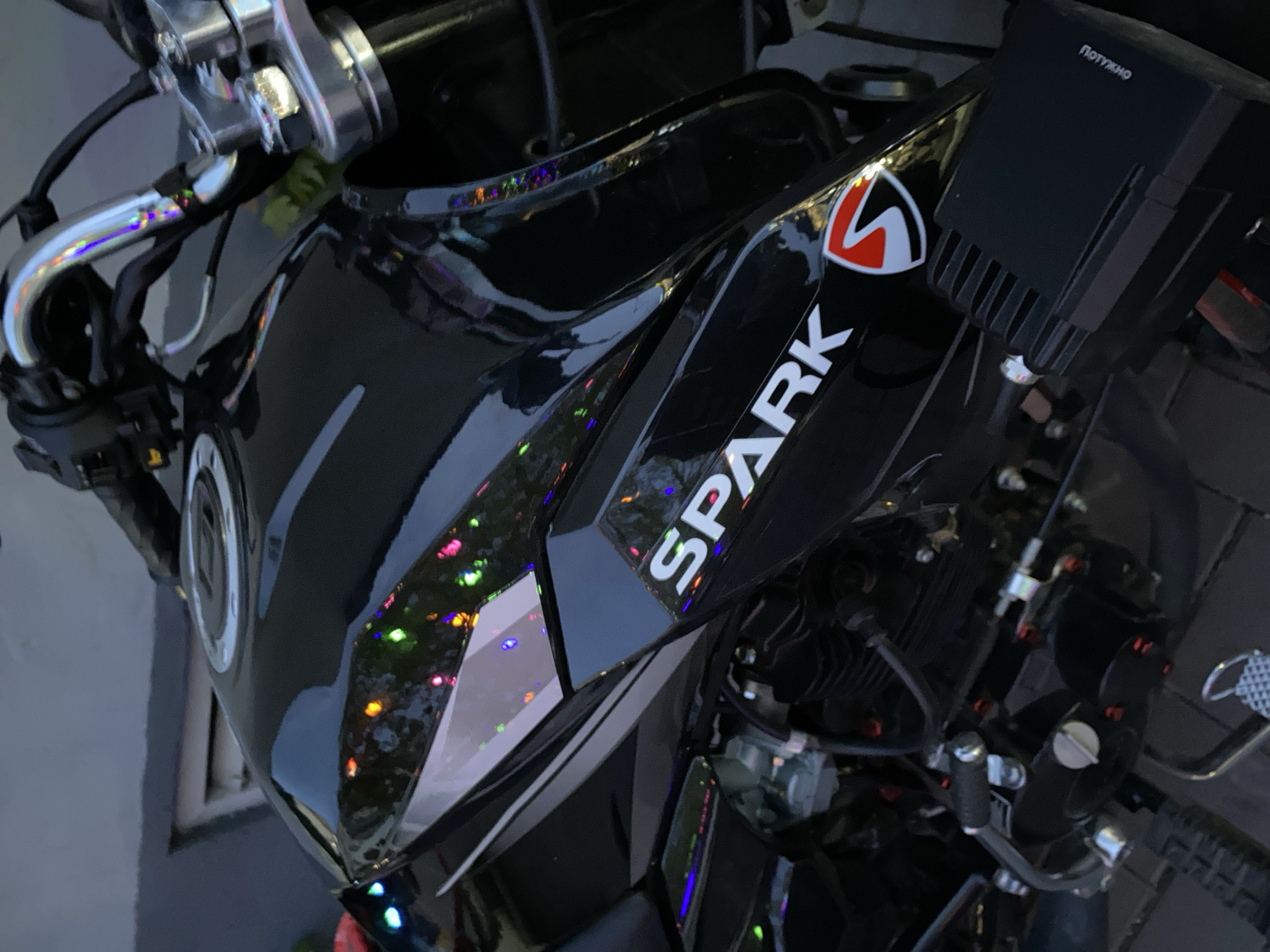 An overhead close-up shot captures a glossy black motorcycle, likely stationary. The prominent features include the fuel tank, handlebars, and a portion of the engine. The fuel tank is emblazoned with the white text "SPARK" and an accompanying red and white stylized logo. A chrome fuel cap is visible on the tank. The motorcycle's highly reflective surface is adorned with numerous small, out-of-focus, colorful light reflections (red, green, blue, yellow), suggesting the presence of decorative lights or a brightly lit environment at night or indoors. Near the front, a black, rectangular electronic device, possibly an auxiliary light or control unit, is mounted, bearing the white Cyrillic text "ПОТУЖНО" (meaning "POWERFUL"). Metallic handlebars and levers are visible to the left. Parts of the engine block are visible to the right, featuring some red-colored bolts. The background is dark and indistinct, indicating a low-light setting.
