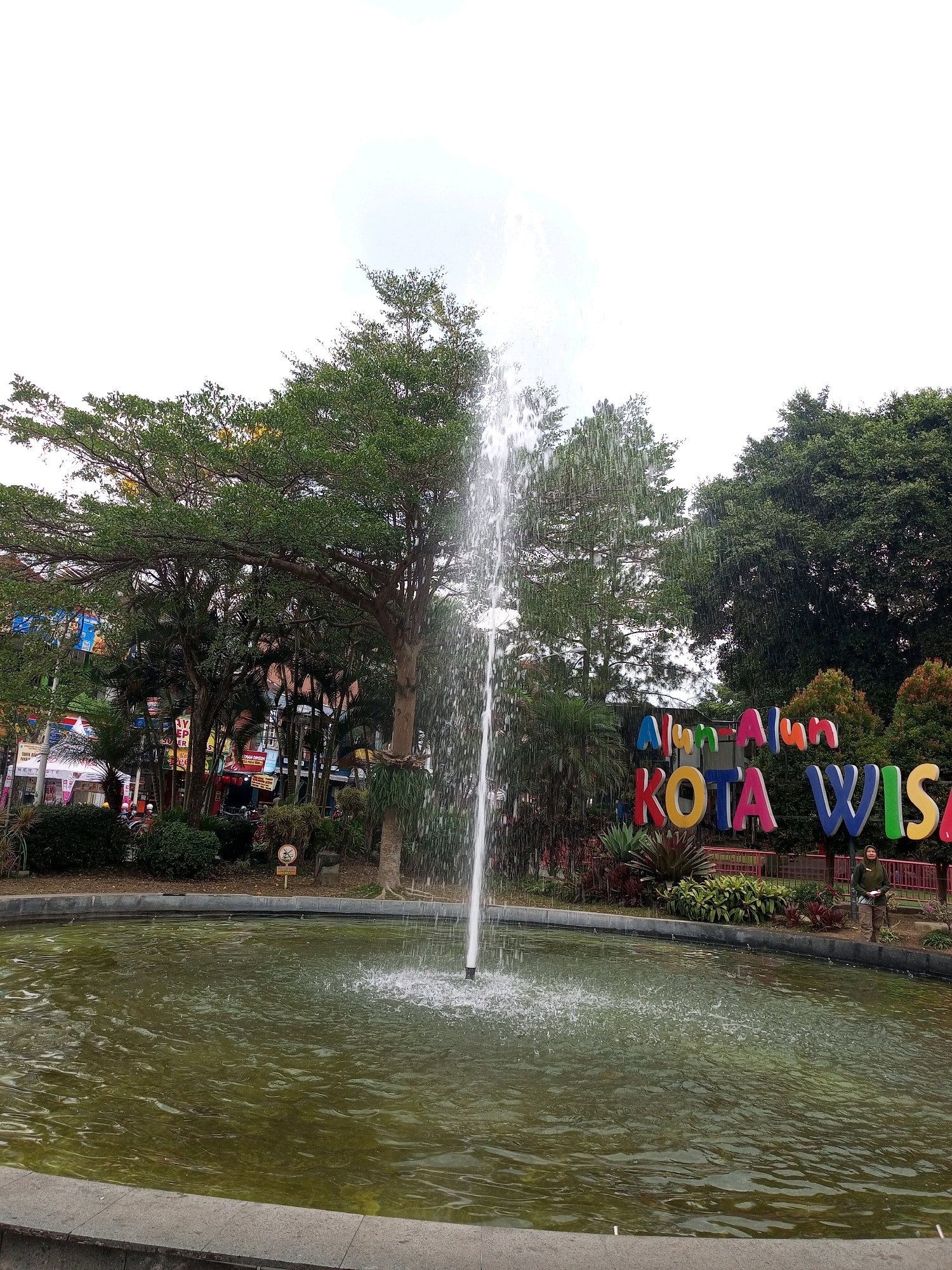 The image depicts a public fountain in front of a large sign that reads "Alun-Alun Kota Wisata" in colorful, three-dimensional letters. The fountain is actively spraying water upwards, creating a misty effect. Behind the fountain, there is lush greenery, including several large trees with dense foliage and some smaller plants. To the right of the sign, a person wearing a green jacket and beige pants stands near the edge of the fountain. The "Alun-Alun Kota Wisata" sign, also known as the Batu City Square, is a popular tourist attraction in Batu, Indonesia, often featuring vibrant decorations and recreational facilities. The overall scene suggests a pleasant outdoor environment, likely a park or town square, with a focus on recreational and tourist appeal.