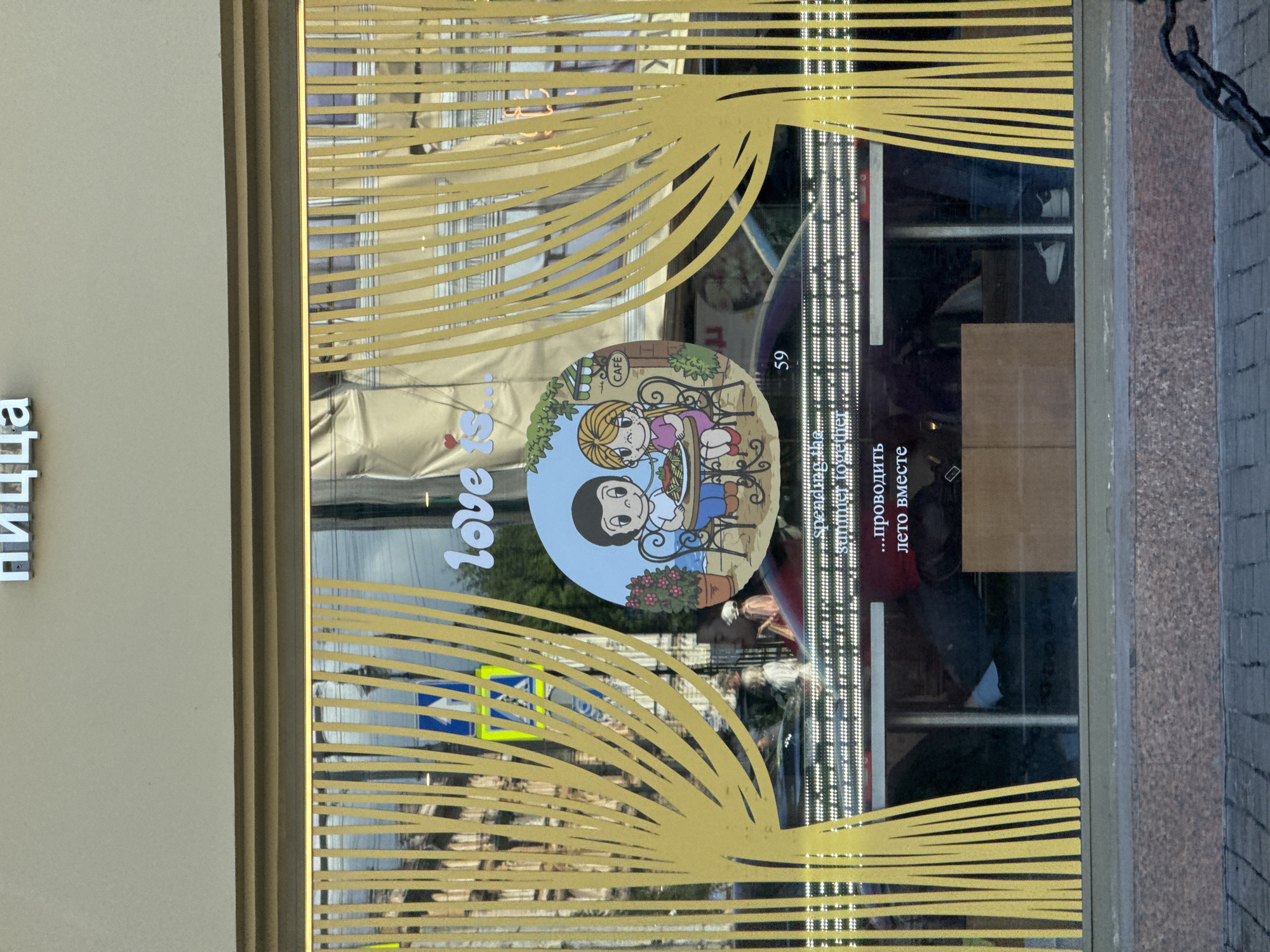 The image shows a storefront window of a cafe named "Love is...". The window is decorated with yellow vertical stripes that resemble noodles, and a circular graphic featuring characters from the "Love is..." comic strip. A boy and a girl are depicted sitting at a table outside a cafe, eating. The words "Love is..." are written in white lettering on the window. Below the graphic, there is text in both English and Russian. The English text reads "spending the summer together." The Russian text reads "... проводить лето вместе." This translates to "... spending the summer together." The cafe appears to be located on a street in Saint Petersburg, Russia, as indicated by the Cyrillic text on the signage. The street is paved with cobblestones, and there is a traffic light visible in the reflection on the window. The interior of the cafe is visible through the window, showing tables and chairs, as well as some people.