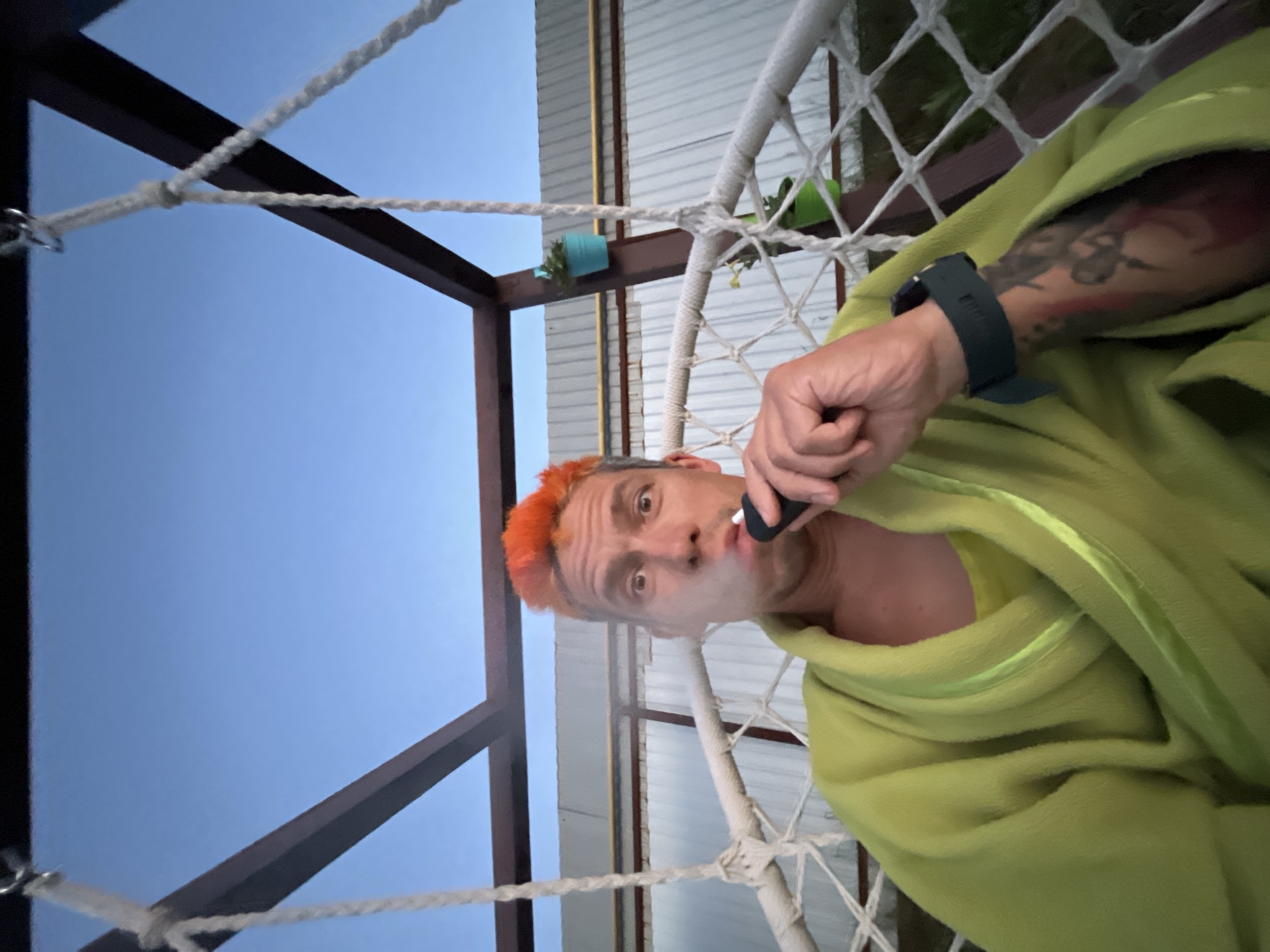 A man with brightly dyed orange hair is sitting in a hammock-like chair, exhaling a puff of smoke from an electronic cigarette. He is wearing a lime green bathrobe over a yellow shirt. His left arm, visible to the viewer, is tattooed. The hammock is suspended from a metal frame. In the background, there is a corrugated metal wall and a clear blue sky. Two small potted plants are attached to one of the metal frame posts. The scene appears to be outdoors, possibly in a backyard or a recreational area, in the city of Chaikovsky, Russia.
